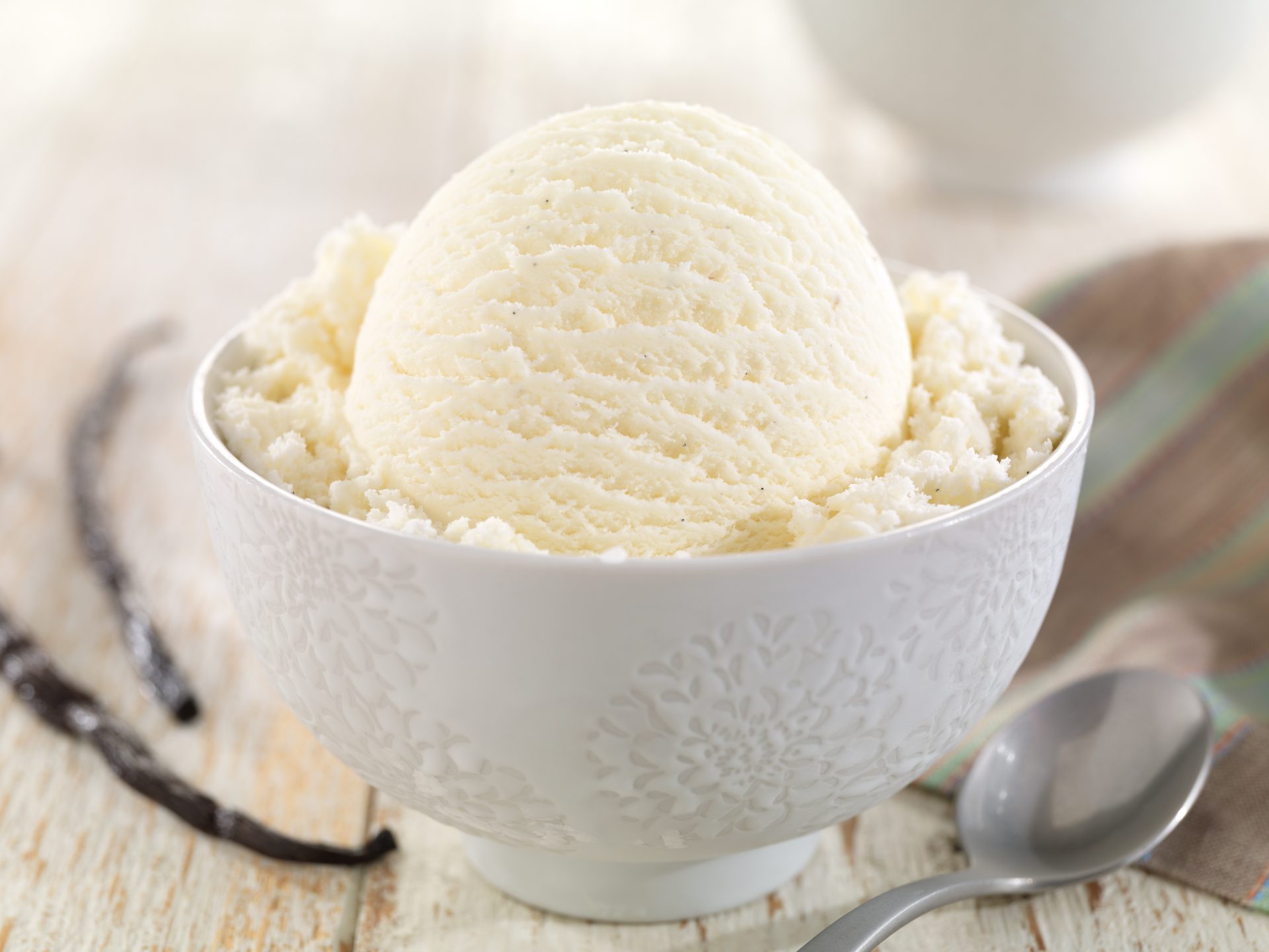 Scoop of vanilla bean ice cream in a white bowl with vanilla pods and a spoon on a light background.