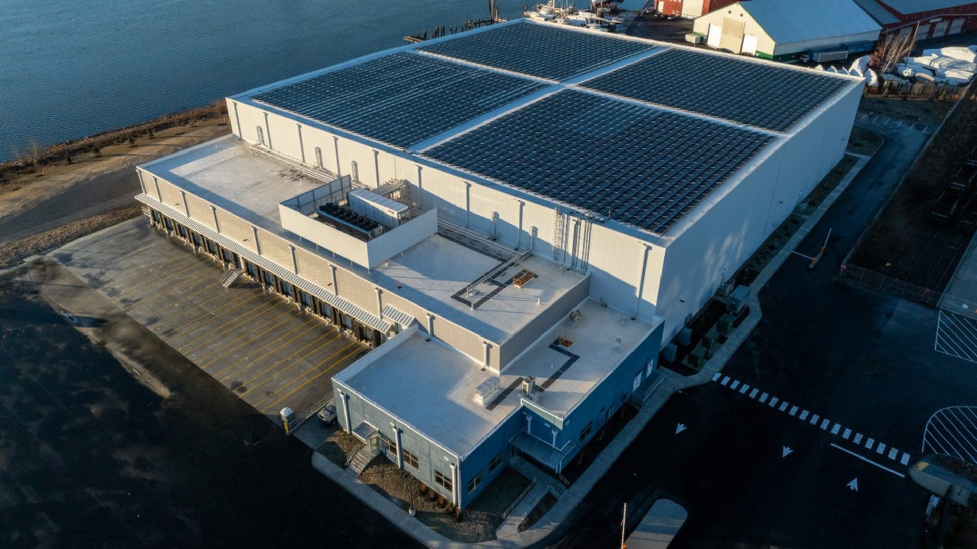Aerial of a large warehouse with a solar-paneled roof, loading docks, and an office, by a river.