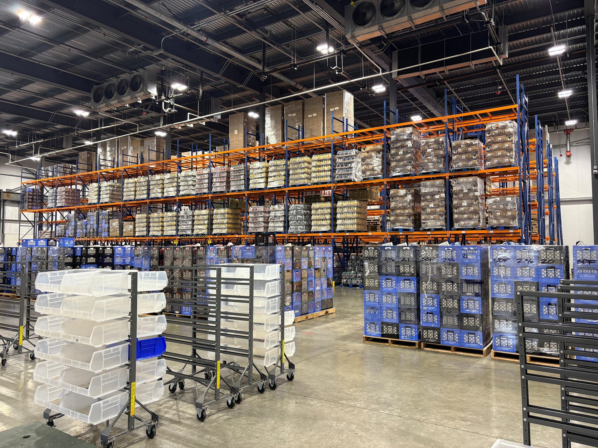 Warehouse interior with tall racks of products, clear bins on carts, and blue crates on pallets.