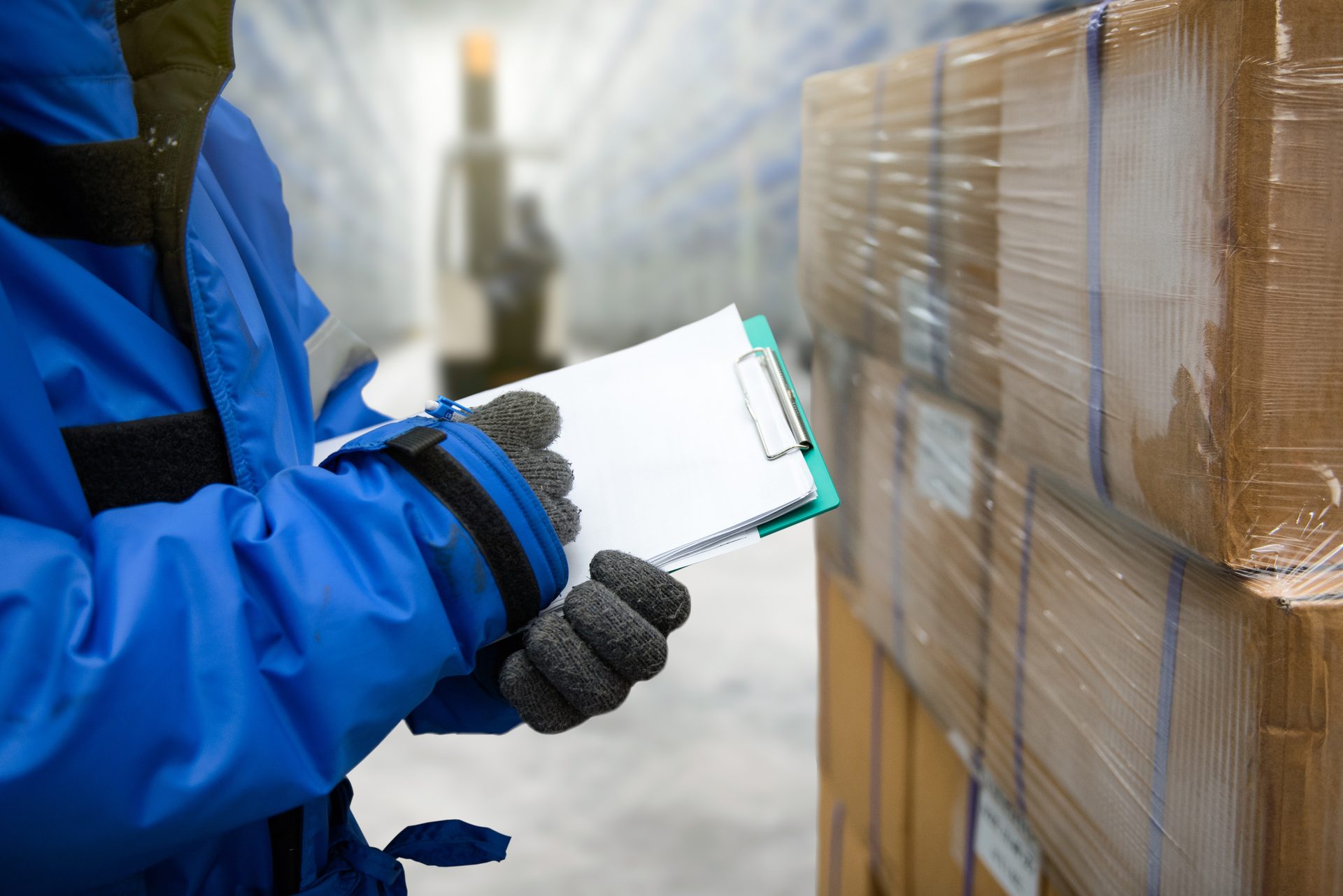 Worker in winter gear holds clipboard, checking boxes in a cold warehouse.