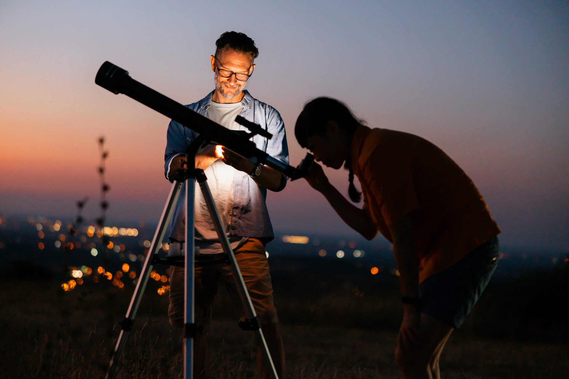 Man and child look through a telescope at dusk, city lights in background.