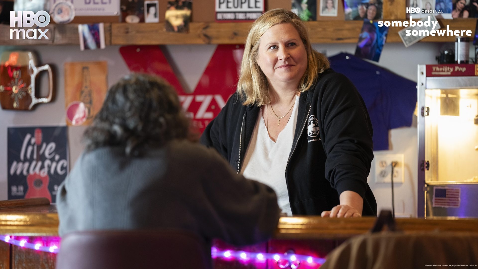 A woman smiles at a bar, facing another person. Promo for HBO Max's "Somebody Somewhere."