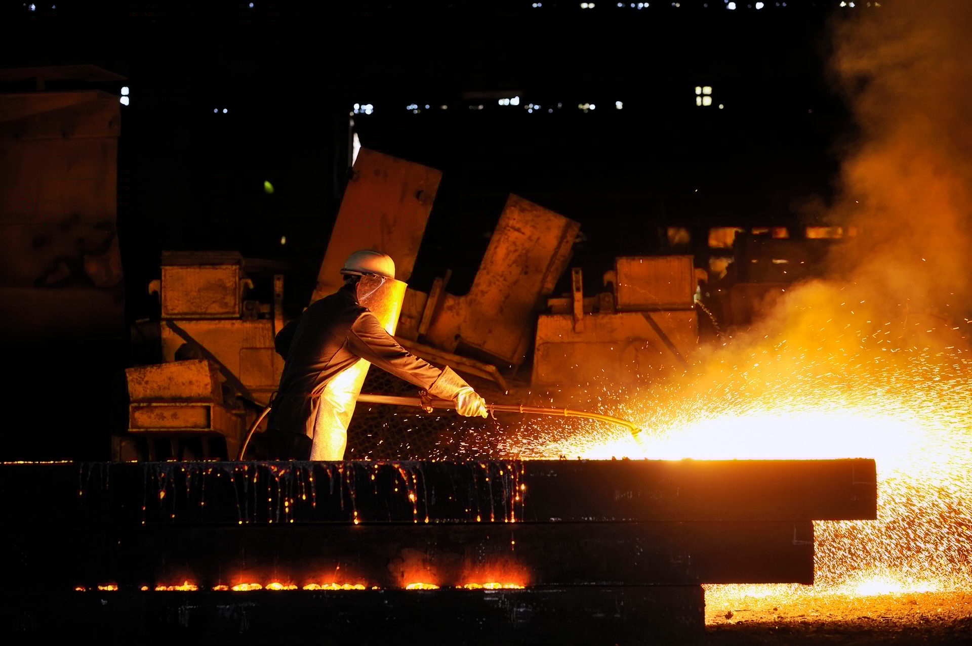 Worker in safety gear cutting metal with torch, intense sparks light up the darkness.