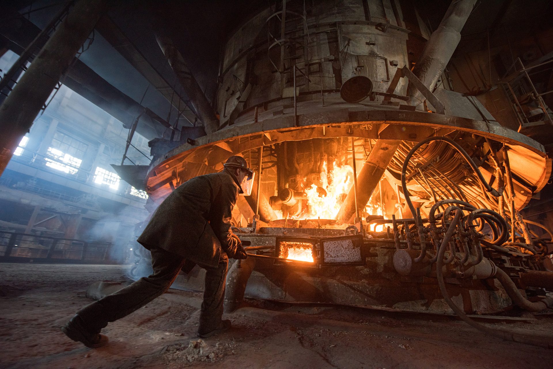 Worker in protective gear tends to a large, fiery furnace in a steel mill.