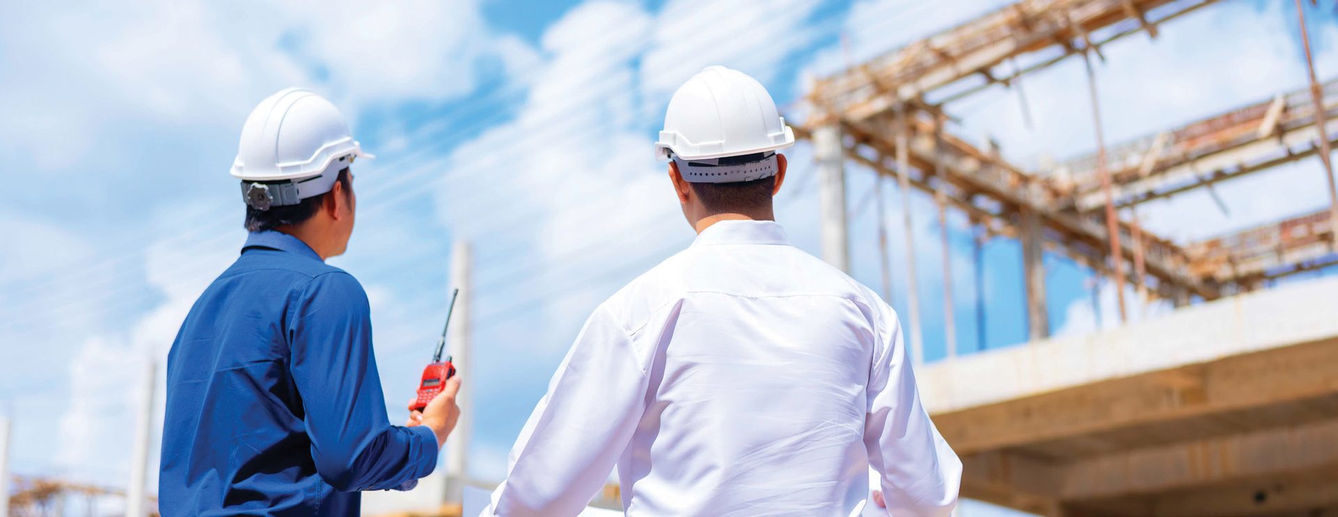 Two construction engineers in hard hats looking at a building structure. One holds a walkie-talkie.