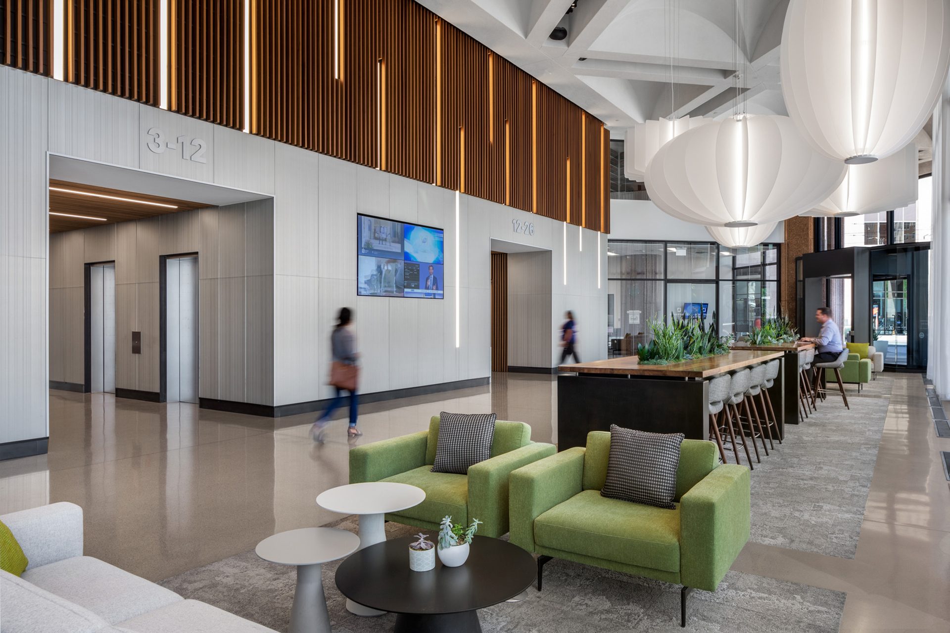 Modern, spacious office lobby with a lounge area, communal bar table, elevators, and decorative lighting.