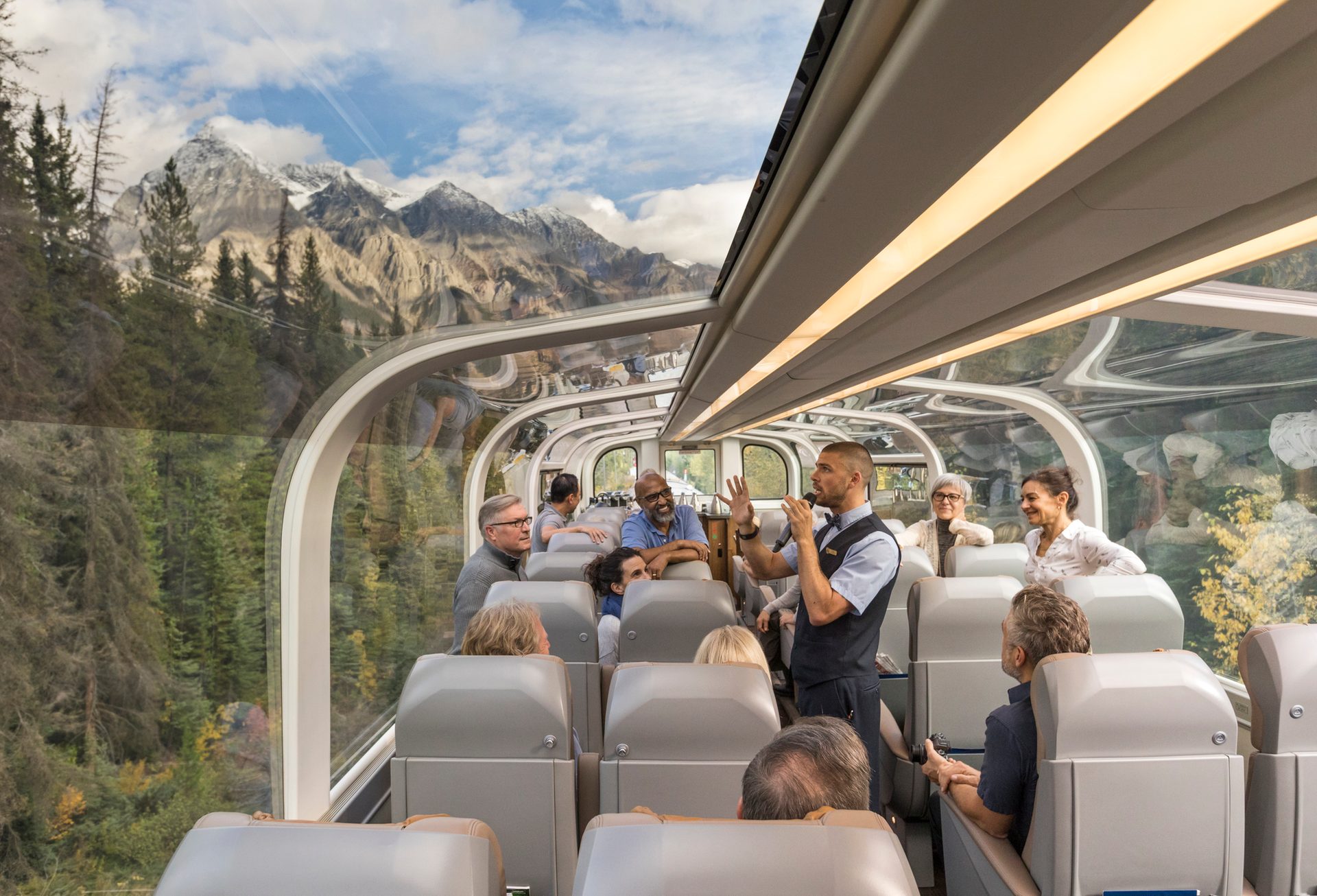 Passengers enjoy mountain views from a glass-domed luxury train while an employee speaks into a microphone.