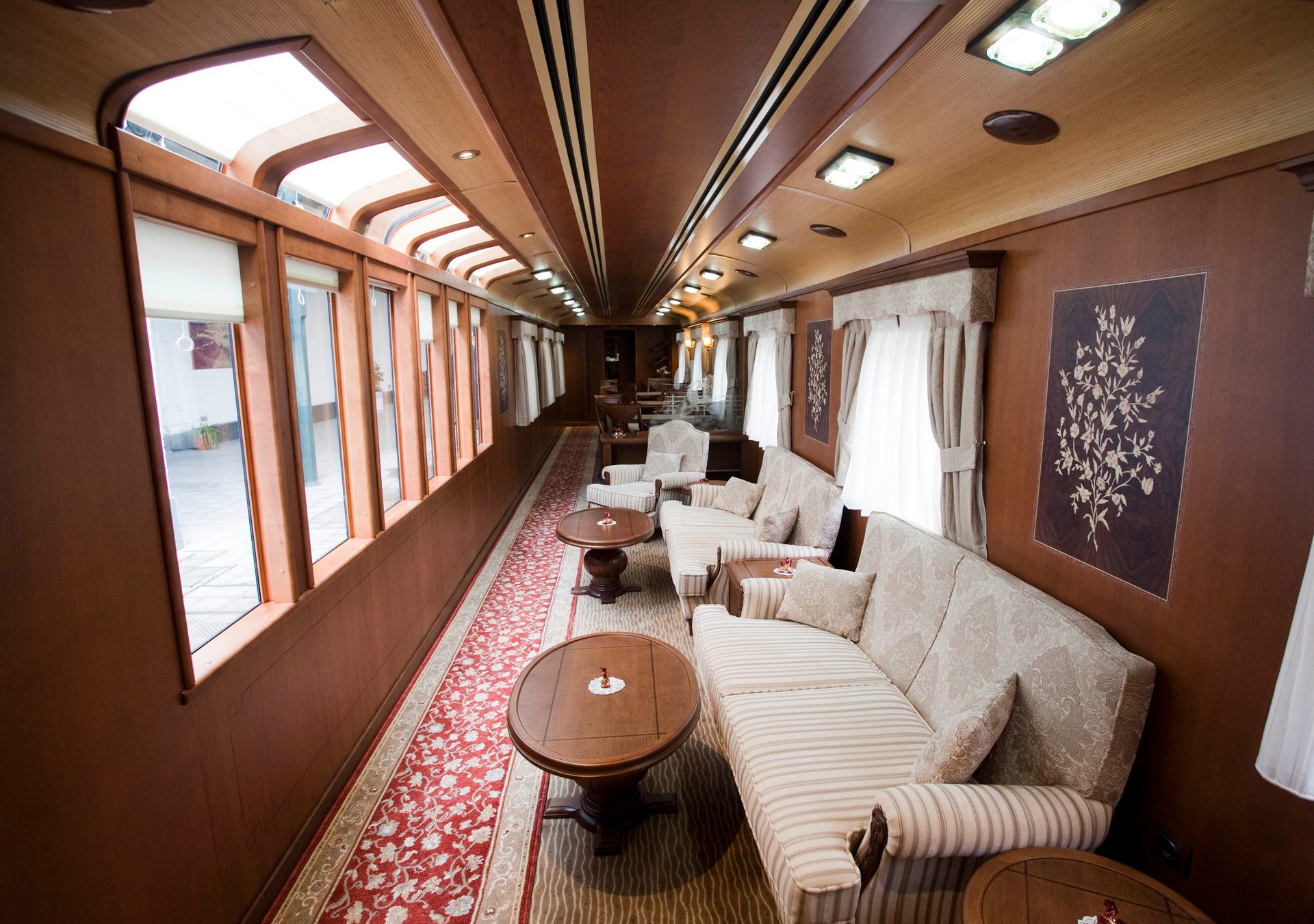 Luxurious train lounge with ornate wood paneling, large windows, plush sofas, and a patterned red carpet.