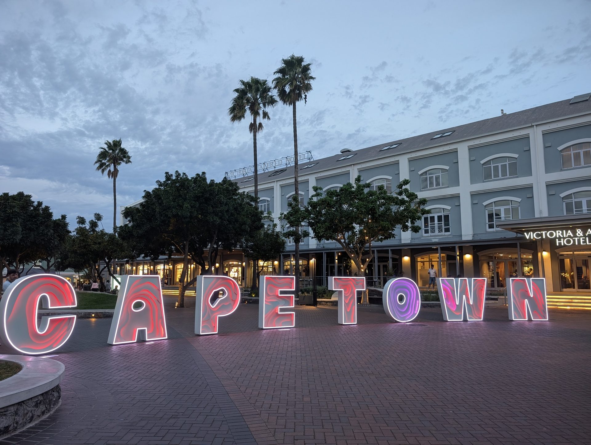 Vibrant CAPETOWN sign glowing at dusk in front of the Victoria & Alfred Hotel and palm trees.