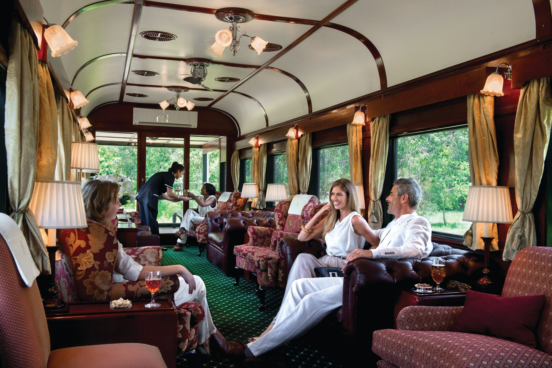Guests relax in a luxurious train carriage with elegant seating, being served drinks by staff.