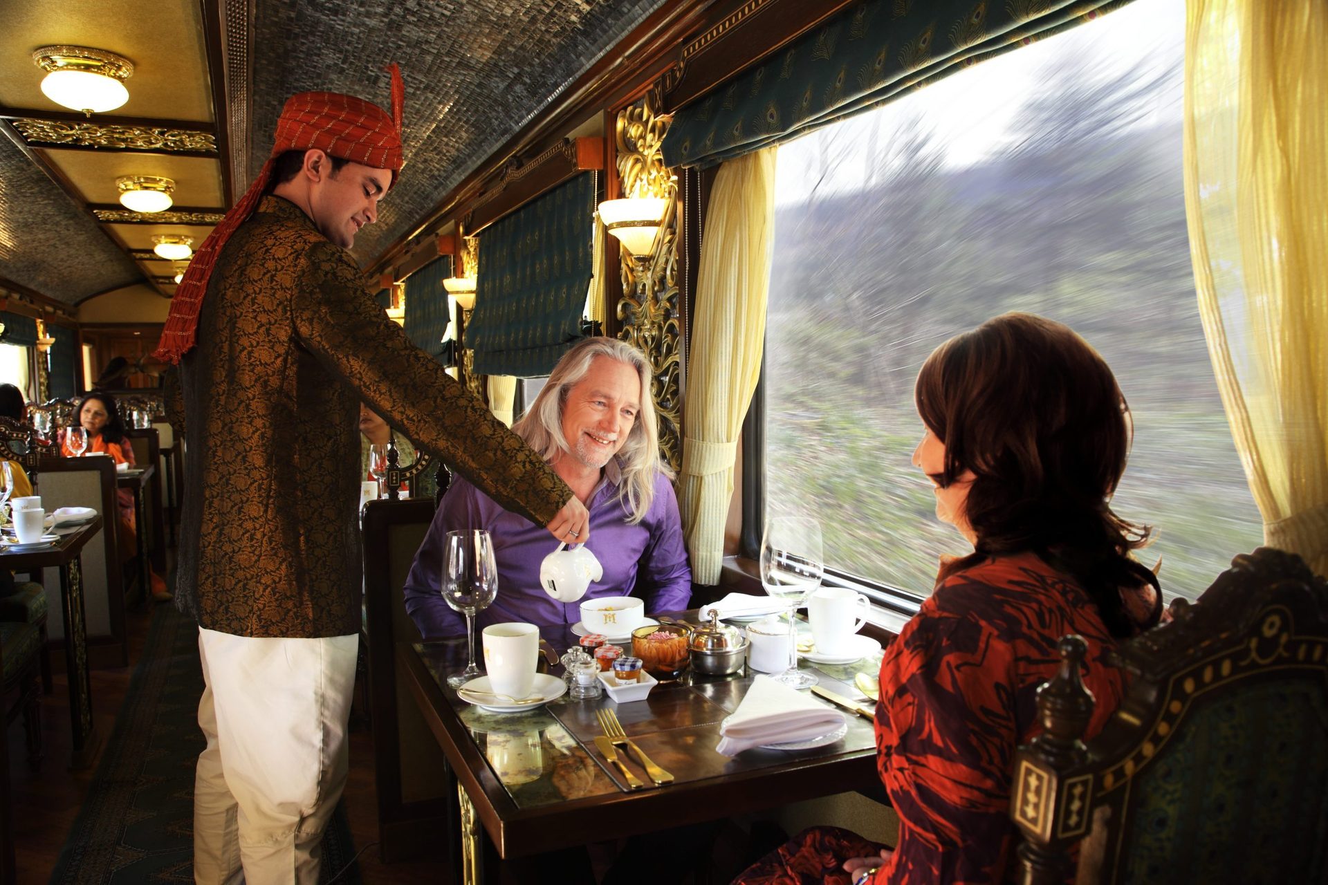 A uniformed server pours tea for two passengers enjoying a meal on a luxurious train journey.