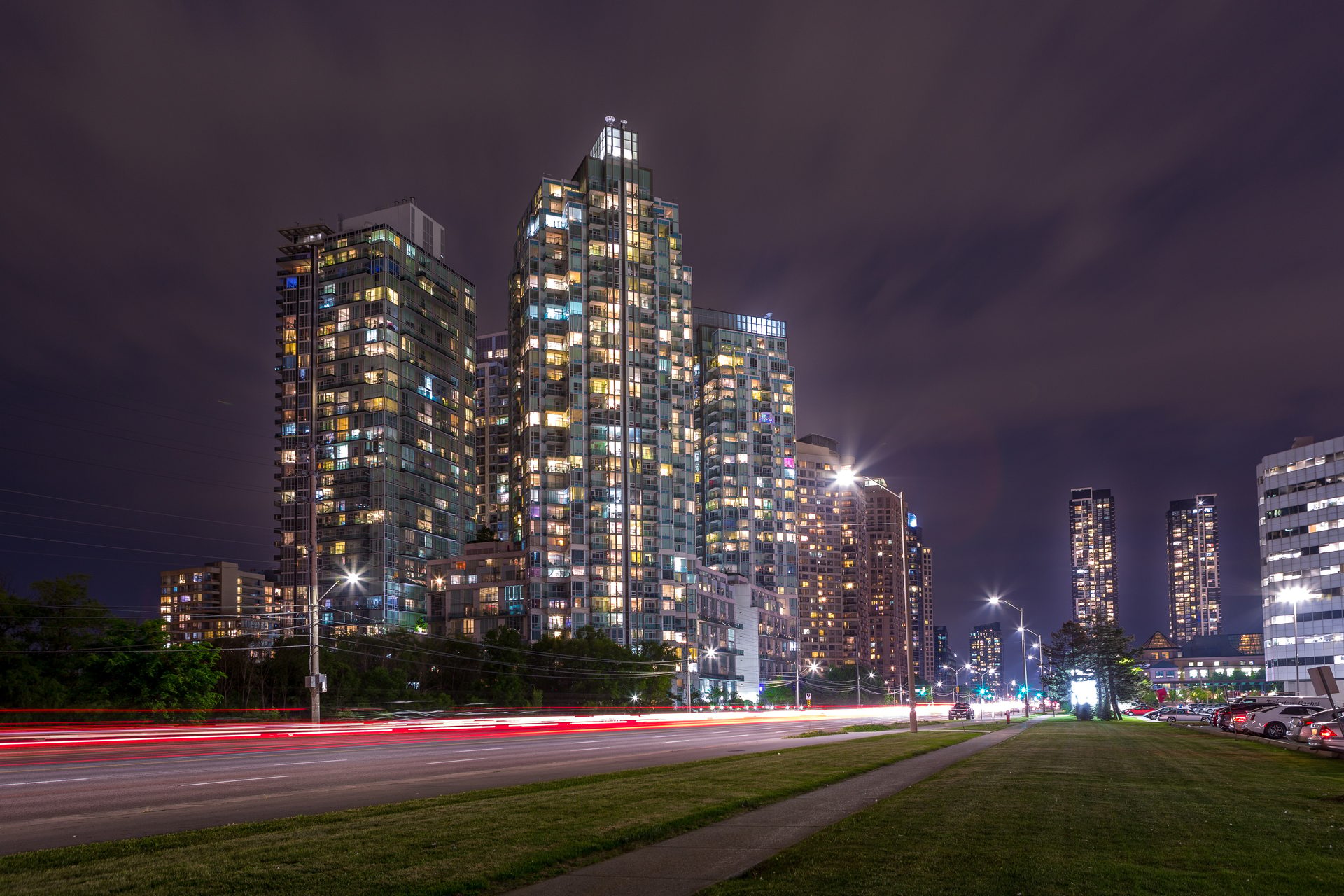 Night cityscape with illuminated skyscrapers, red light trails on a road, and a dark cloudy sky.
