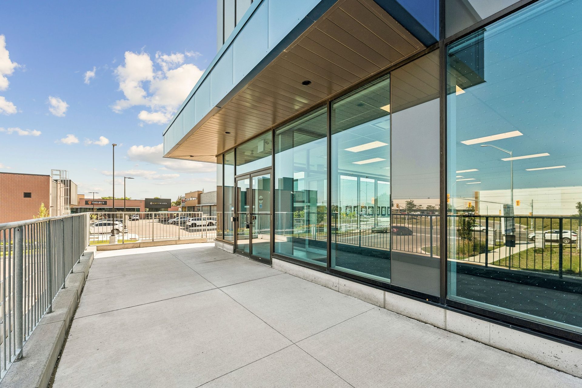 Modern glass building entrance with a spacious concrete patio and railing, leading to a street with distant shops.