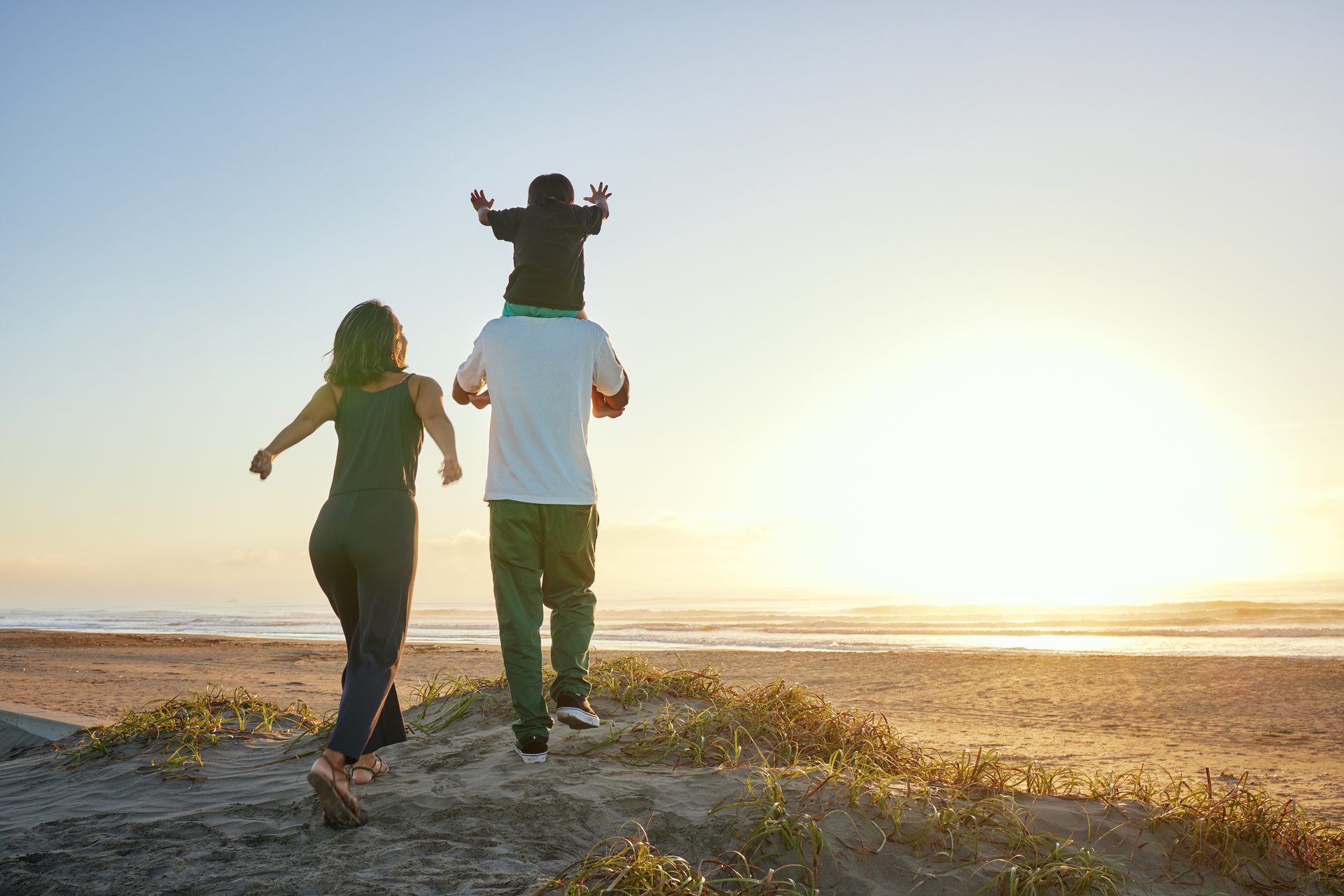 A family with a child on shoulders walks on a beach towards the ocean at sunset.