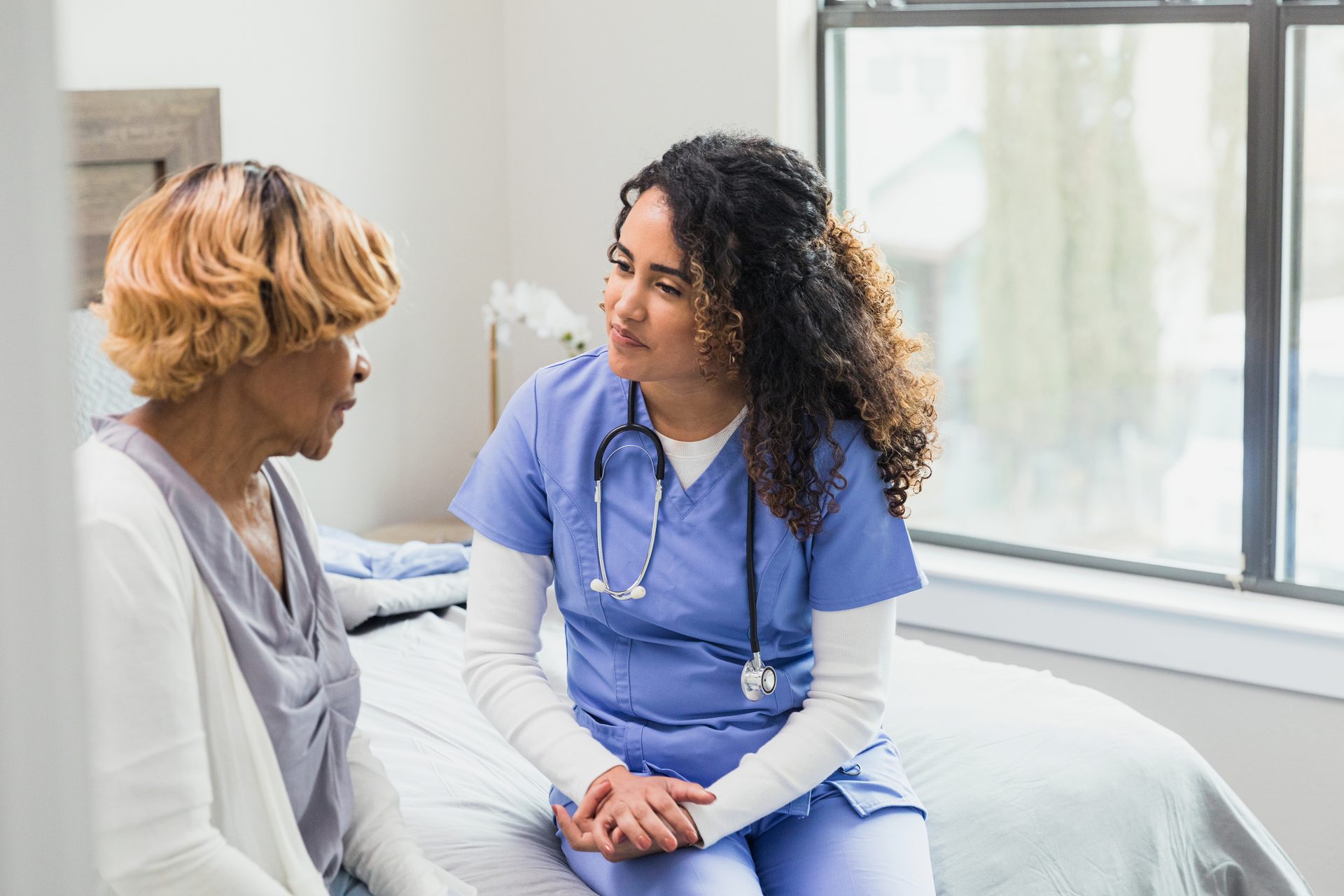 Nurse in blue scrubs and stethoscope attentively talks to an older woman on a bed.