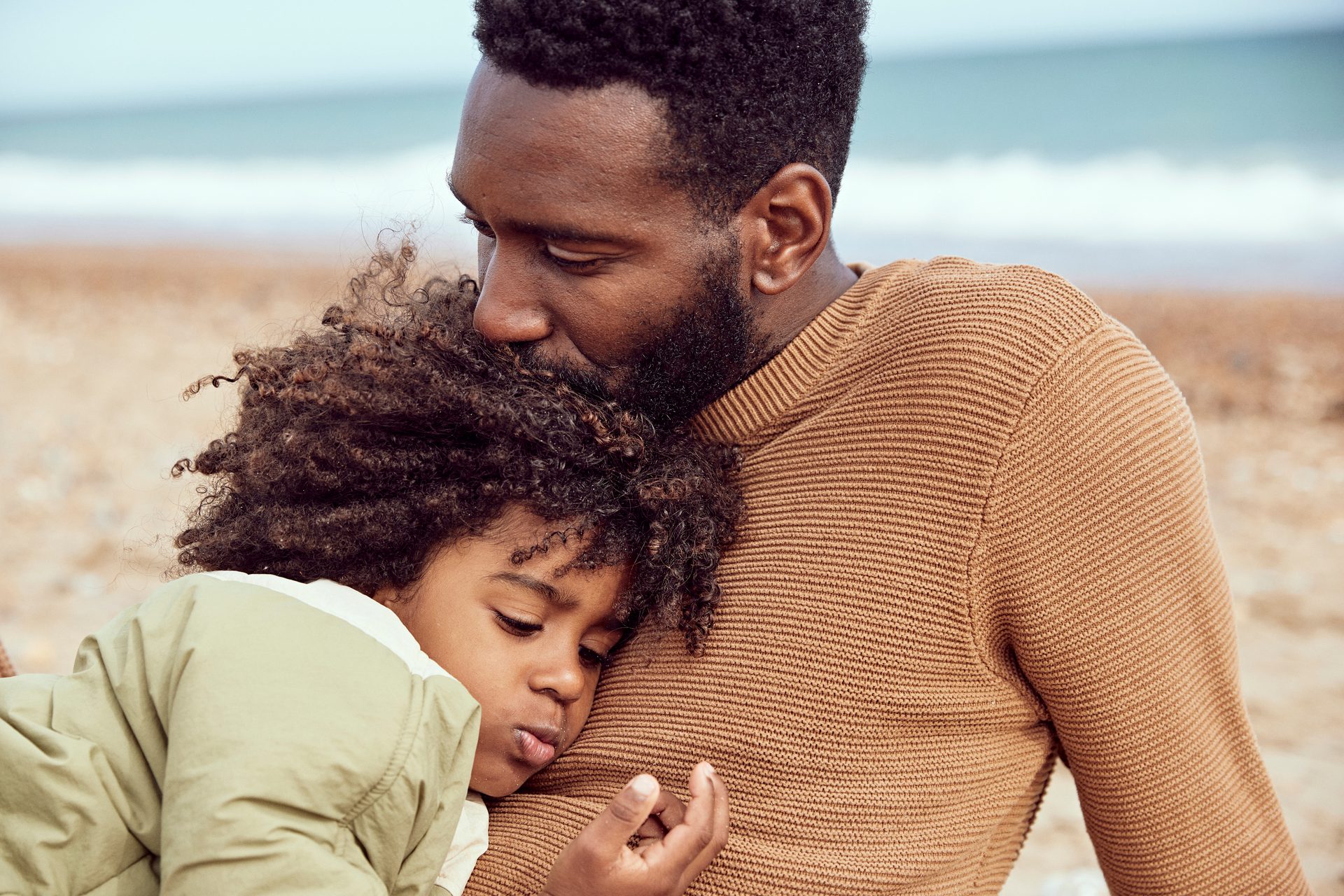 Man kisses child's head on beach; child rests on his chest.