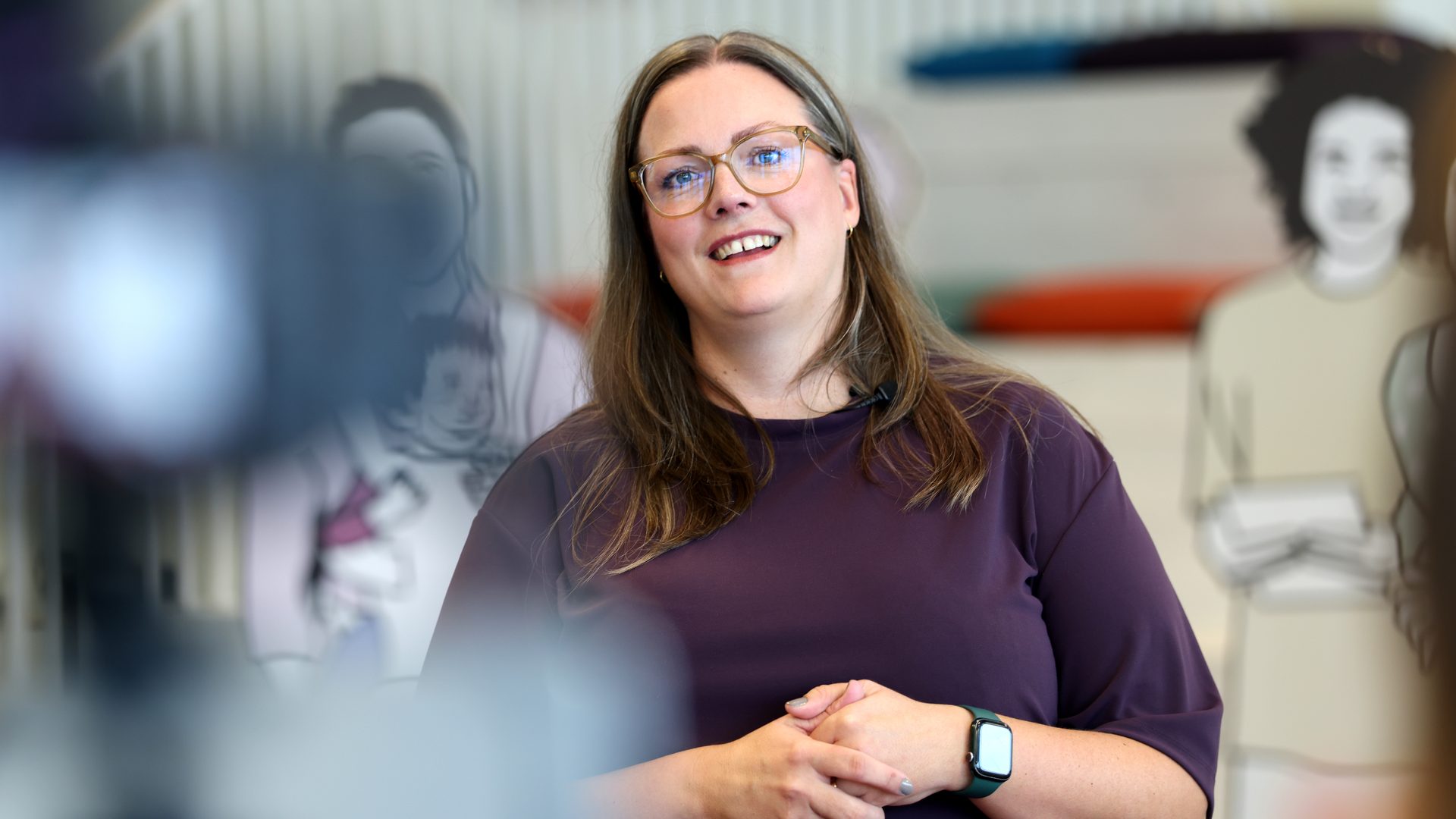 Smiling woman with glasses and long hair in a purple top, with blurred background.