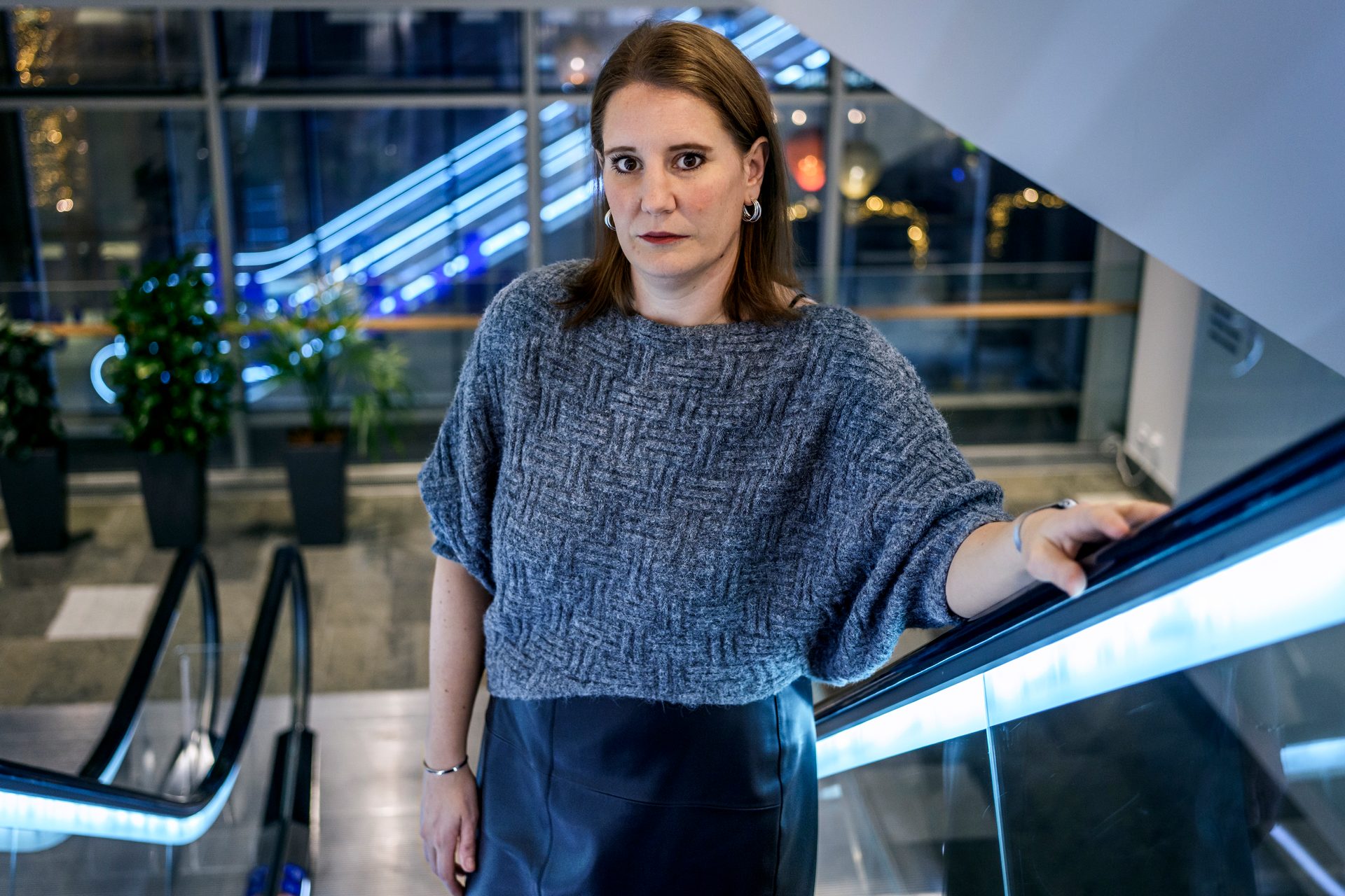 Woman in grey sweater and dark skirt, hand on escalator railing, looking at camera.
