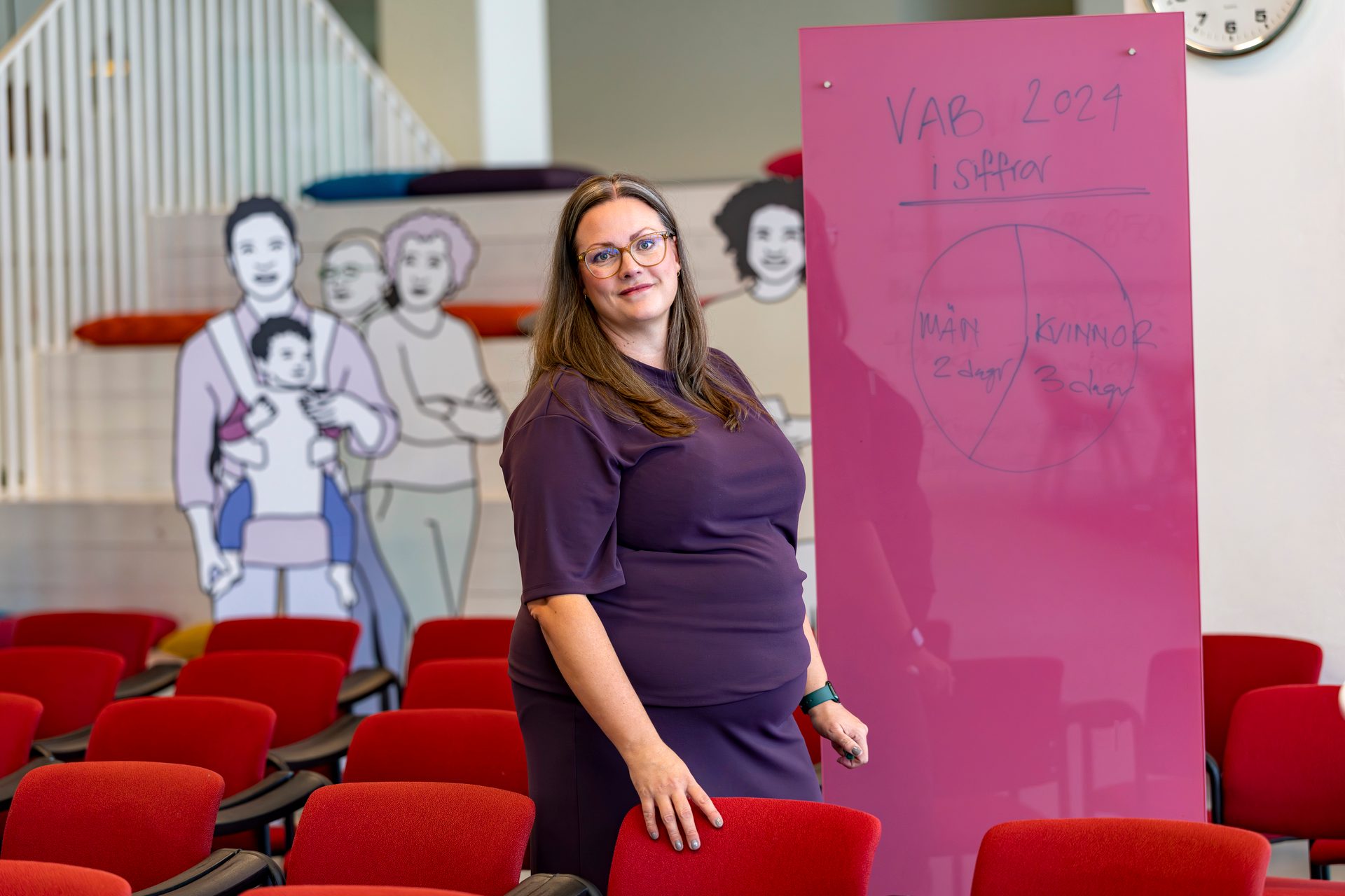 Woman in purple, a pink board with VAB statistics, red chairs, and family figure cutouts.
