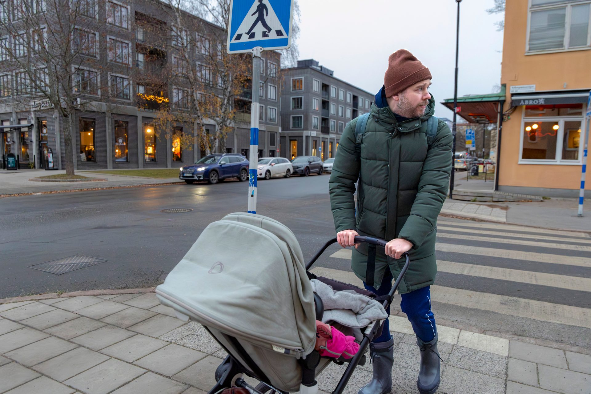 A man in a green puffer jacket and beanie pushes a baby stroller on an urban sidewalk.