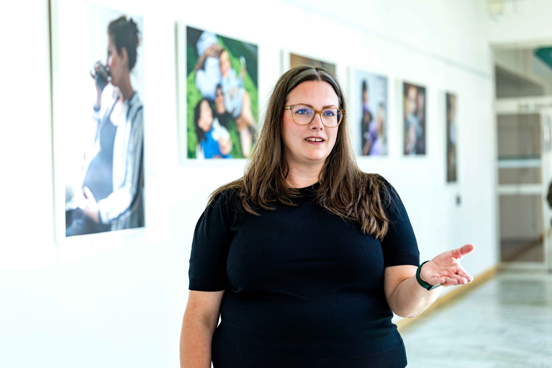 Woman in black shirt, glasses, gesturing in a hallway with photos on a wall.