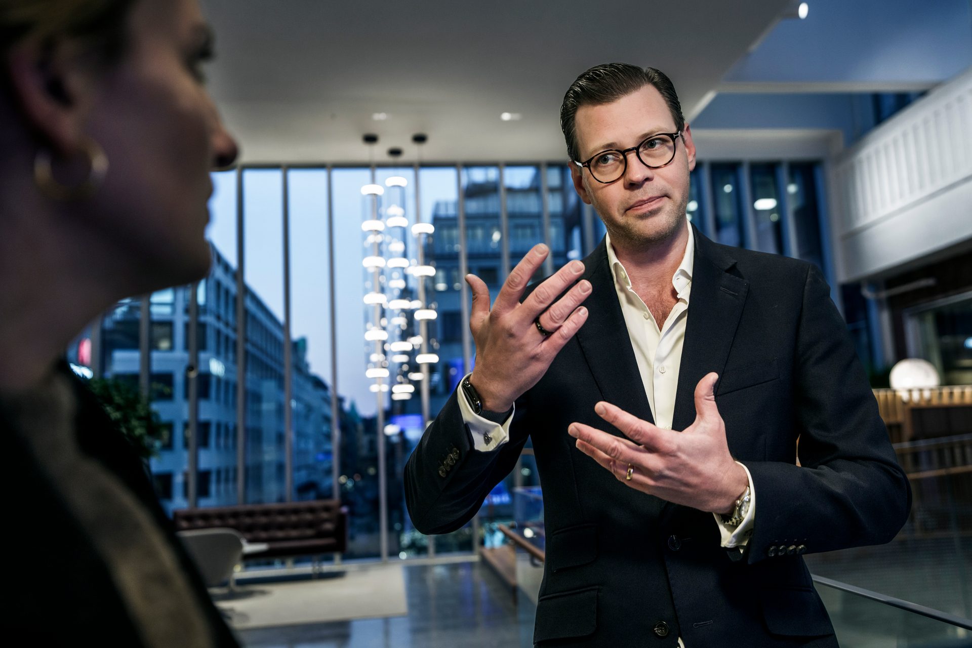 A man in a suit gestures while talking to a blurred woman in a modern interior with a city view.