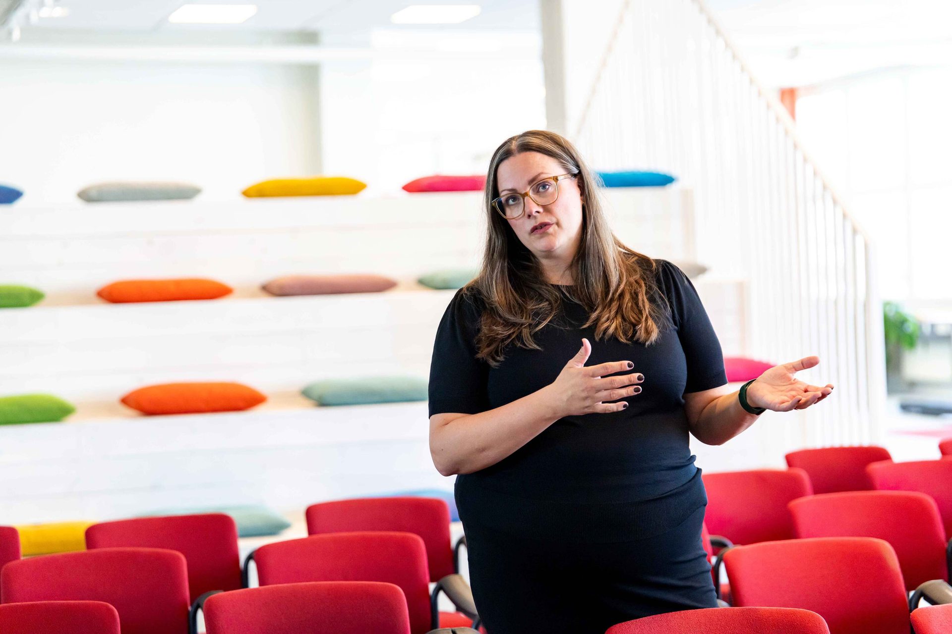 Woman speaking in bright, modern auditorium with colorful tiered seating and red chairs.
