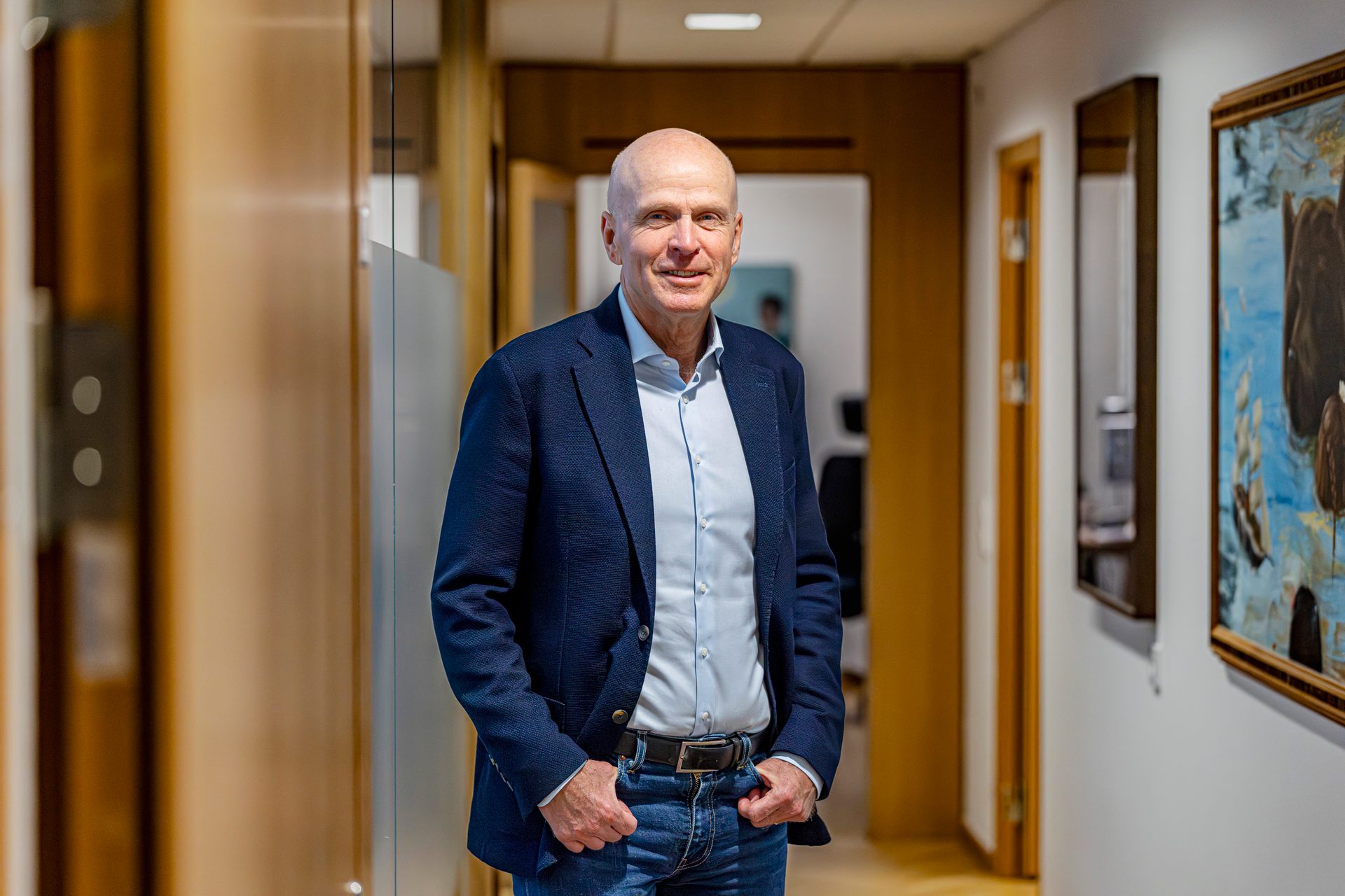 Smiling bald man in a blue blazer and jeans, standing in a hallway.