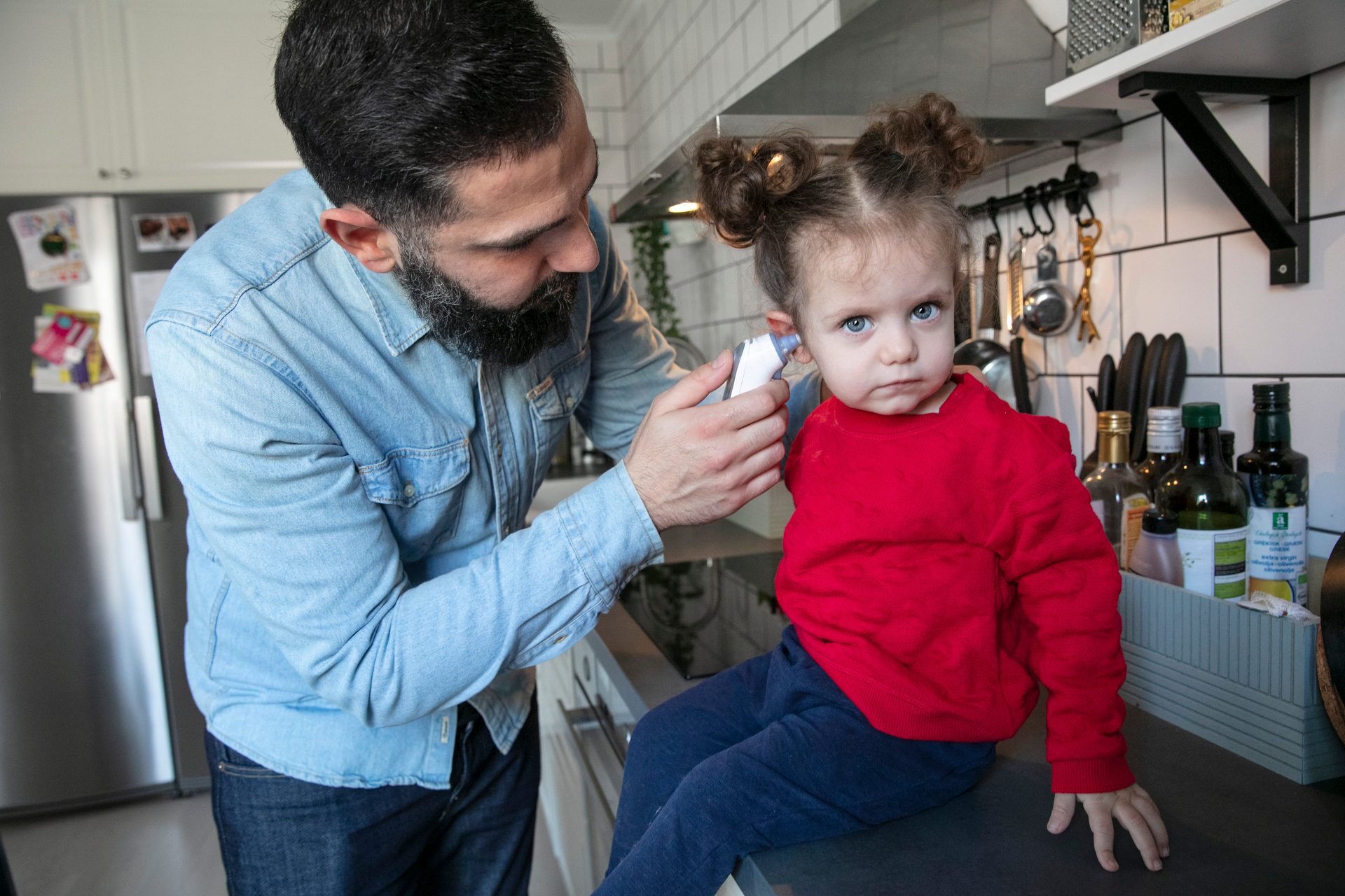 A man takes a serious-looking child's ear temperature in a kitchen.