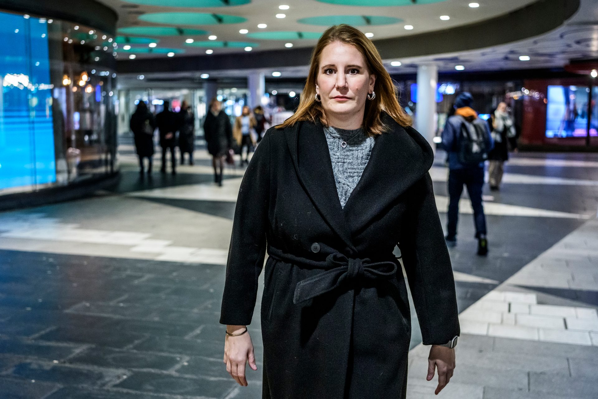 Woman in black coat in a brightly lit indoor public space, other people blurred in the background.