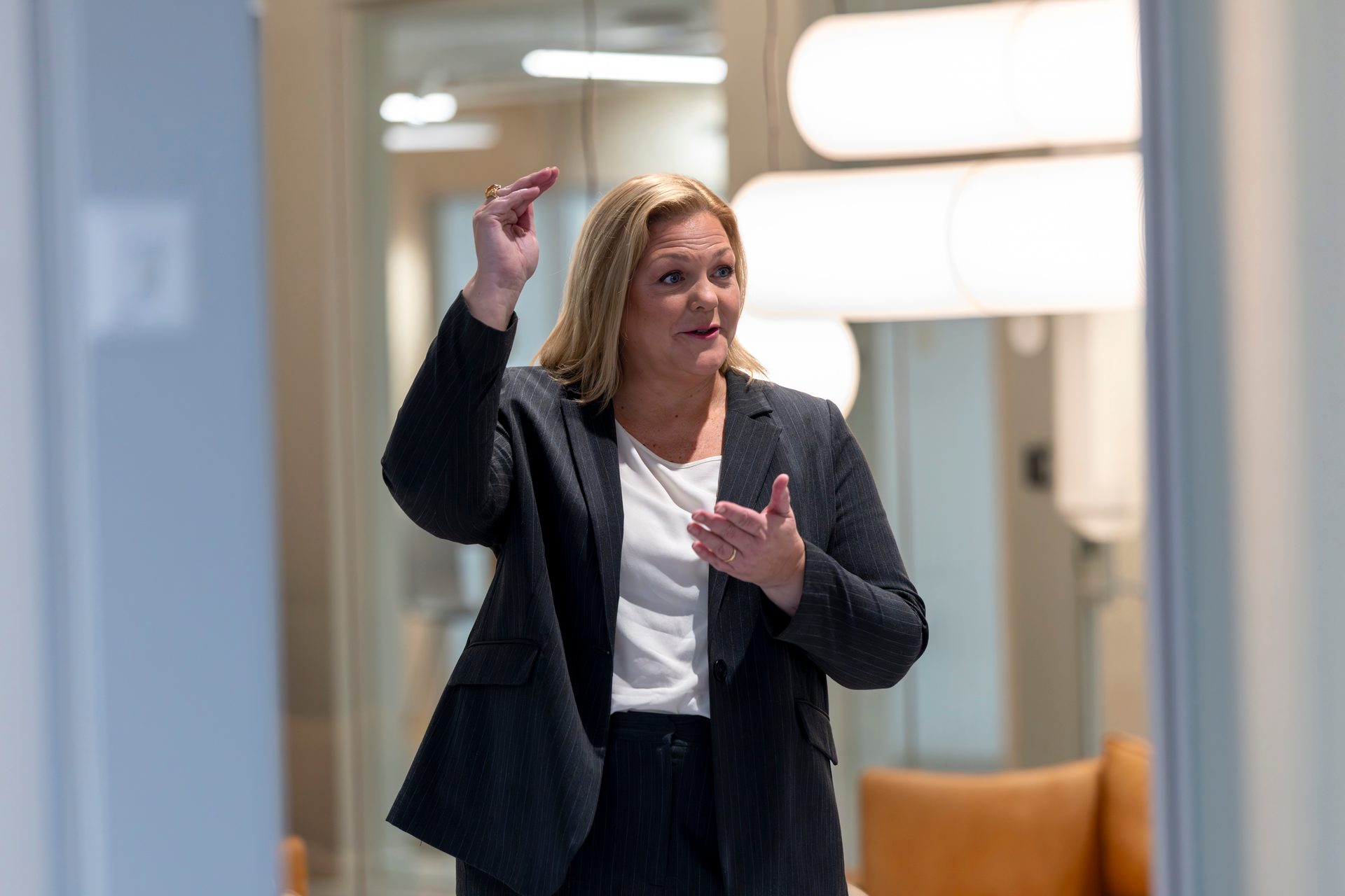 A woman in a pinstripe suit gestures with both hands in a modern office.