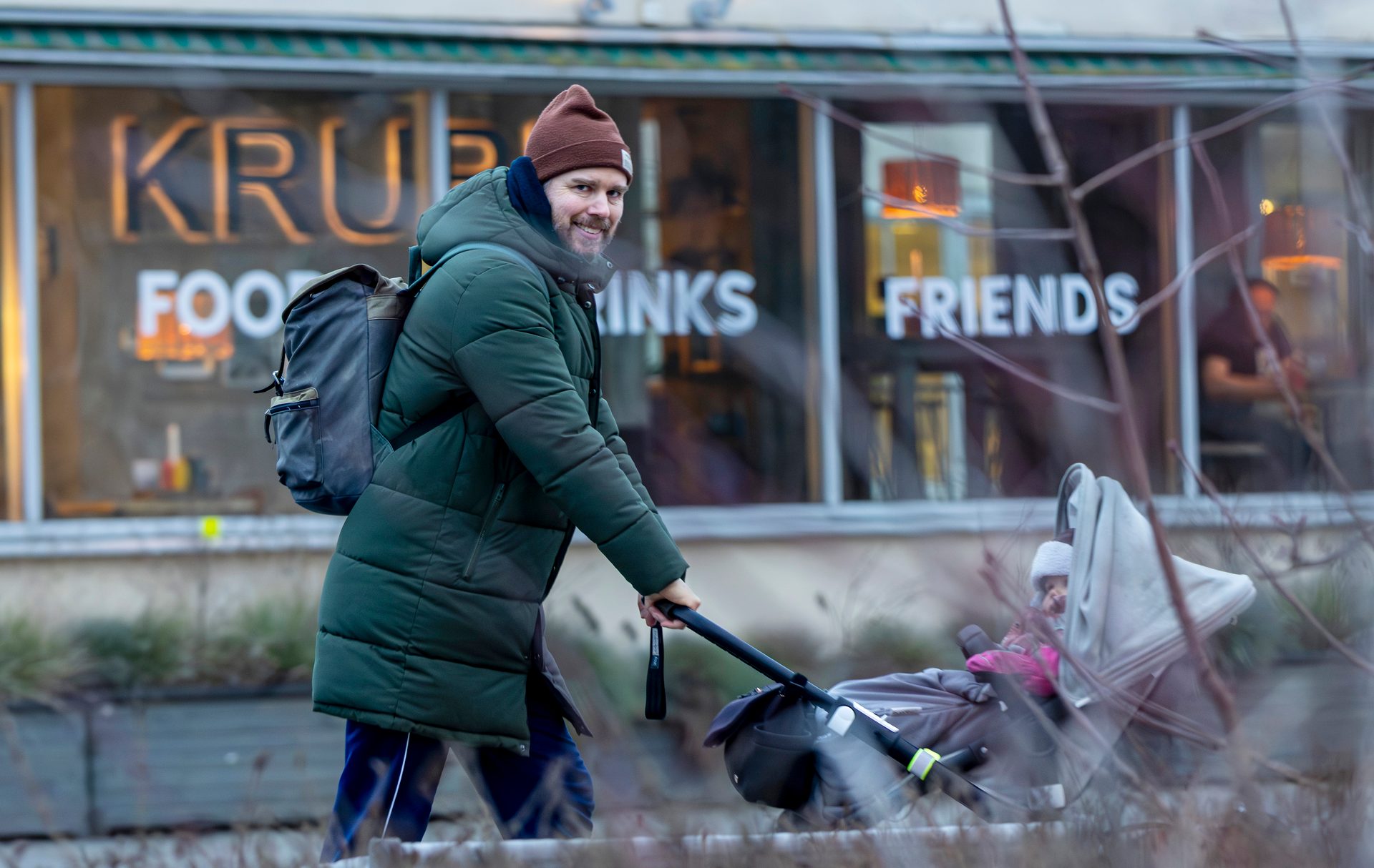 Smiling man in winter coat pushes a stroller with a child past a store advertising "FOOD DRINKS FRIENDS."