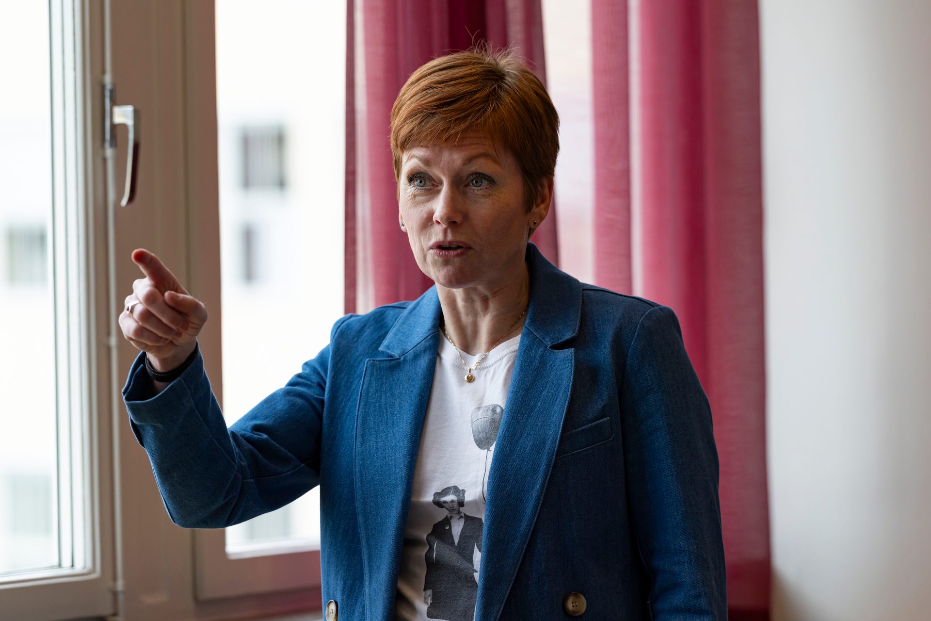 A red-haired woman in a blue jacket points and speaks, with a window and red curtains behind her.