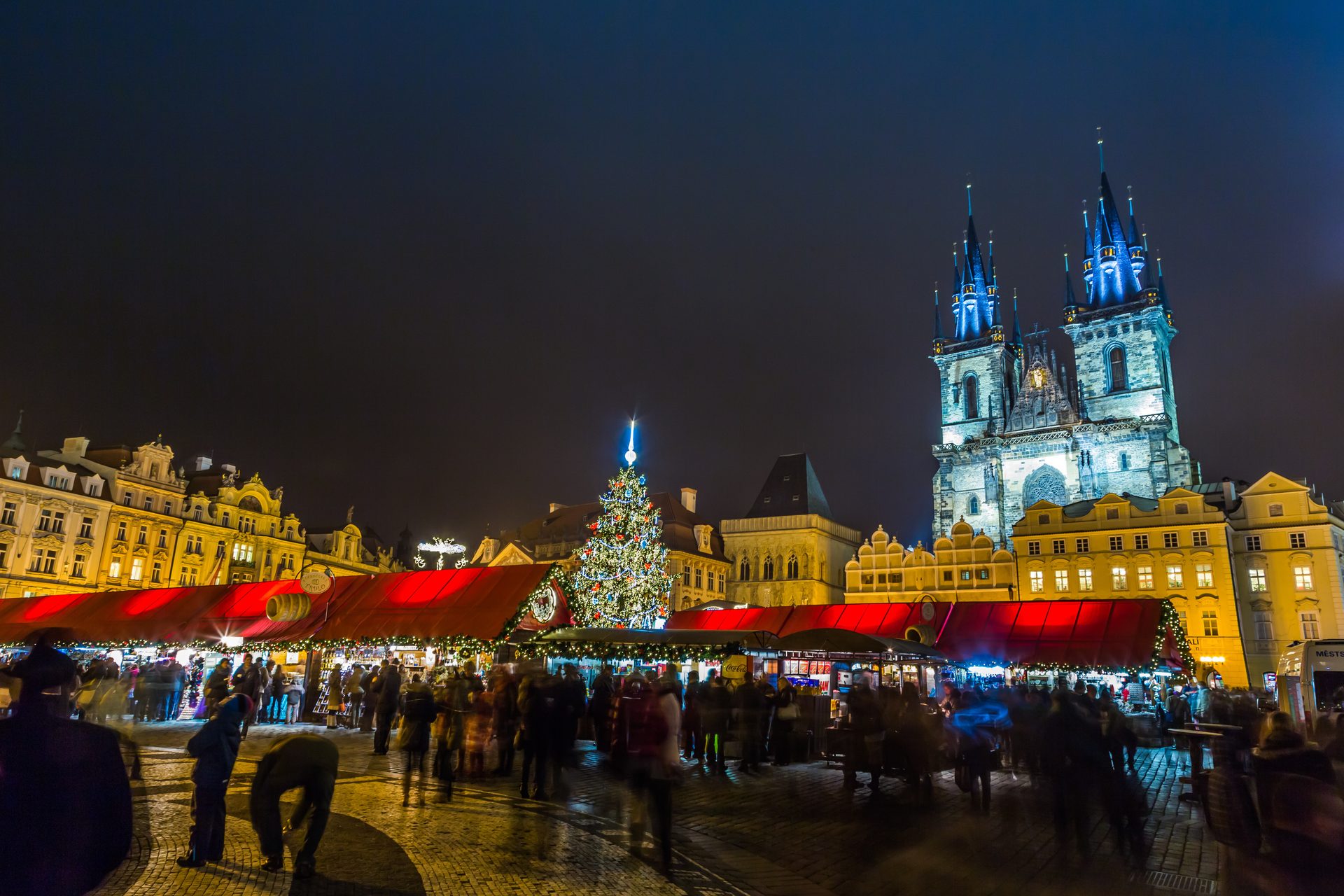 Night Christmas market scene with an illuminated tree, Gothic church, and busy crowds.