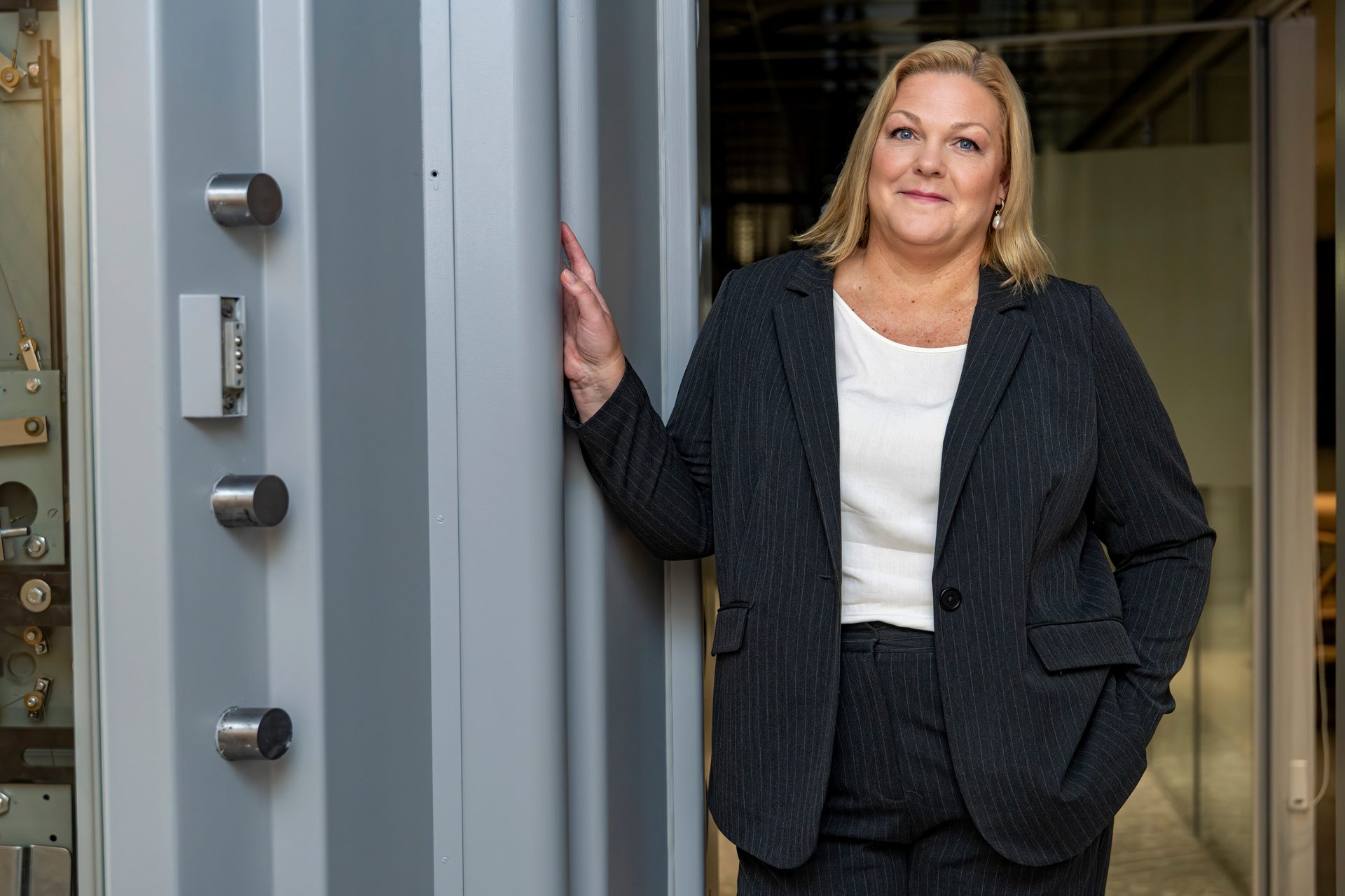 Blonde woman in pinstripe suit, smiling, standing next to an open vault door with exposed locking mechanism.