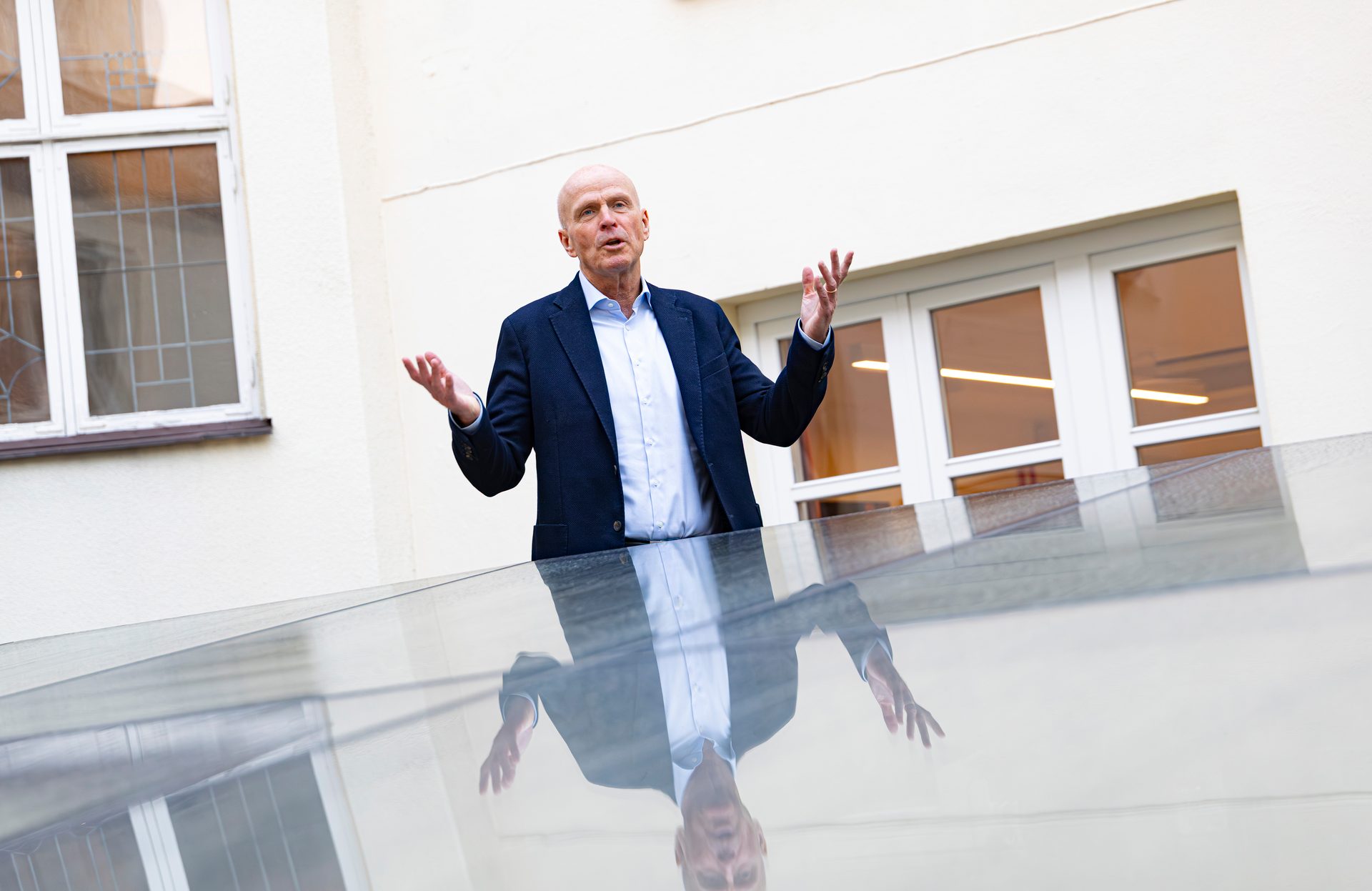 Bald man in a suit gestures, reflected in glass, in front of a building.