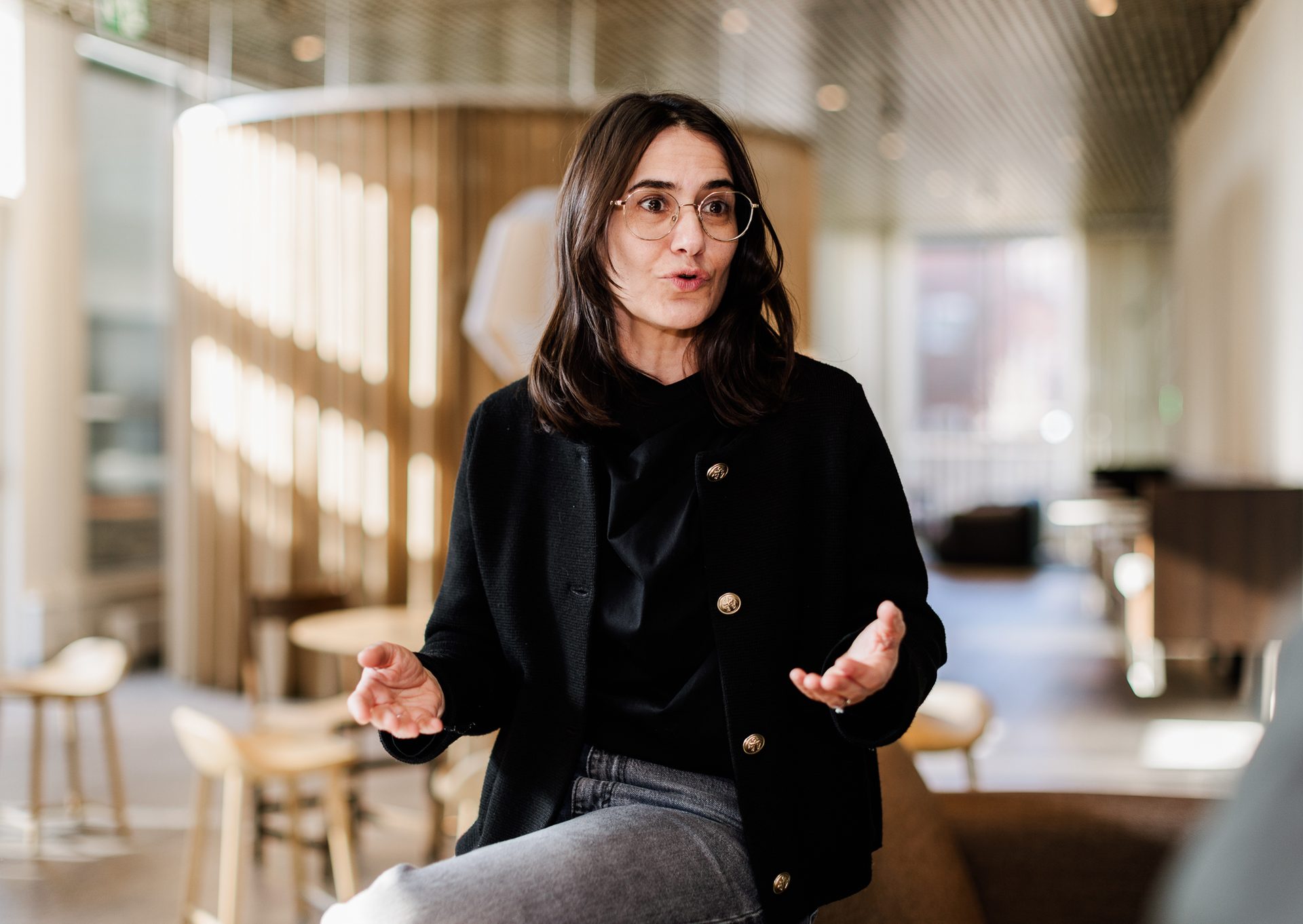 A woman in glasses and a black jacket speaks and gestures with her hands in a modern setting.