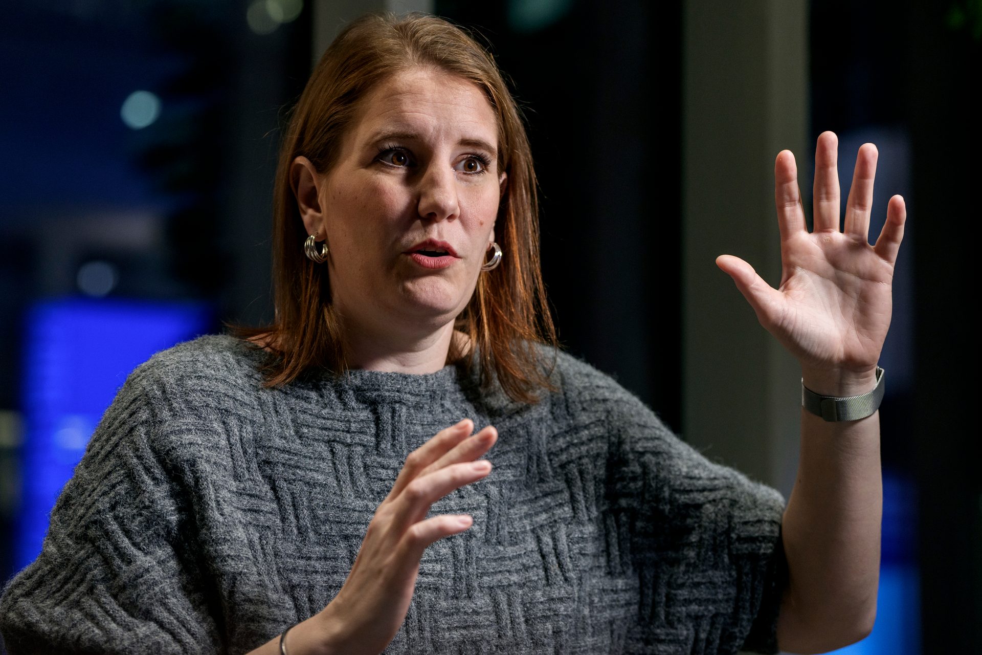 A woman with auburn hair in a textured grey sweater speaks and gestures with both hands.