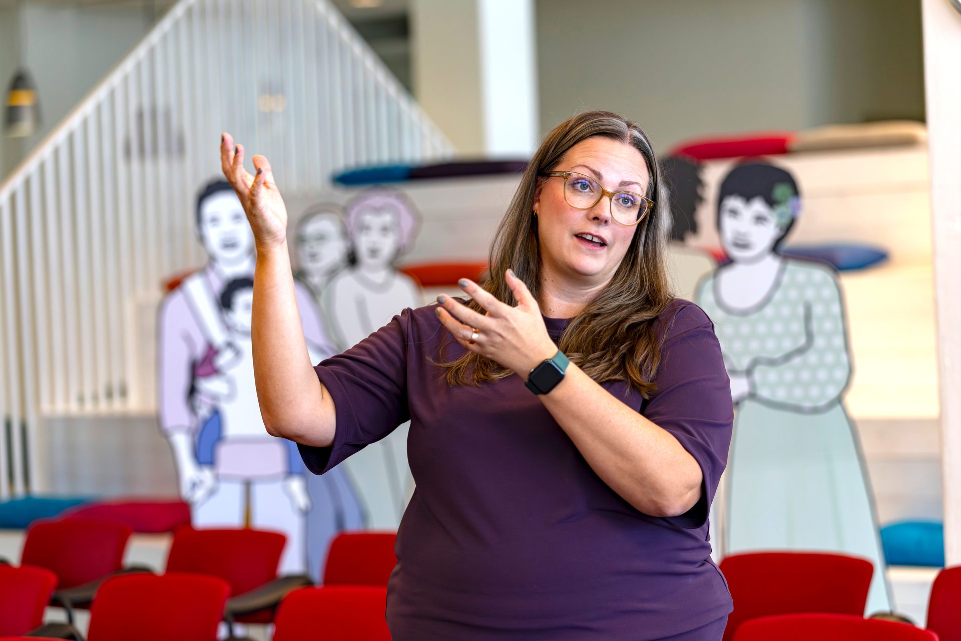 A woman in a purple shirt and glasses gestures animatedly, speaking. Diverse cartoon figures are behind her.