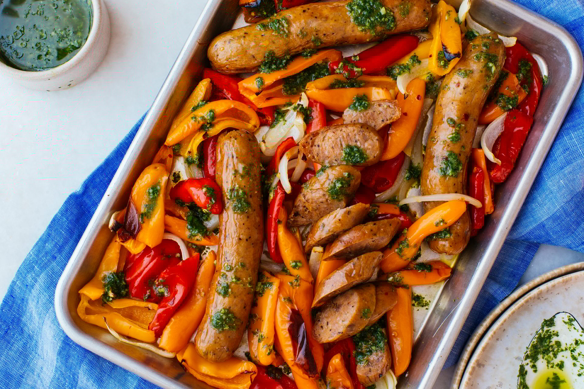 Sheet pan with roasted sausages, red and orange peppers, and onions, drizzled with green sauce.