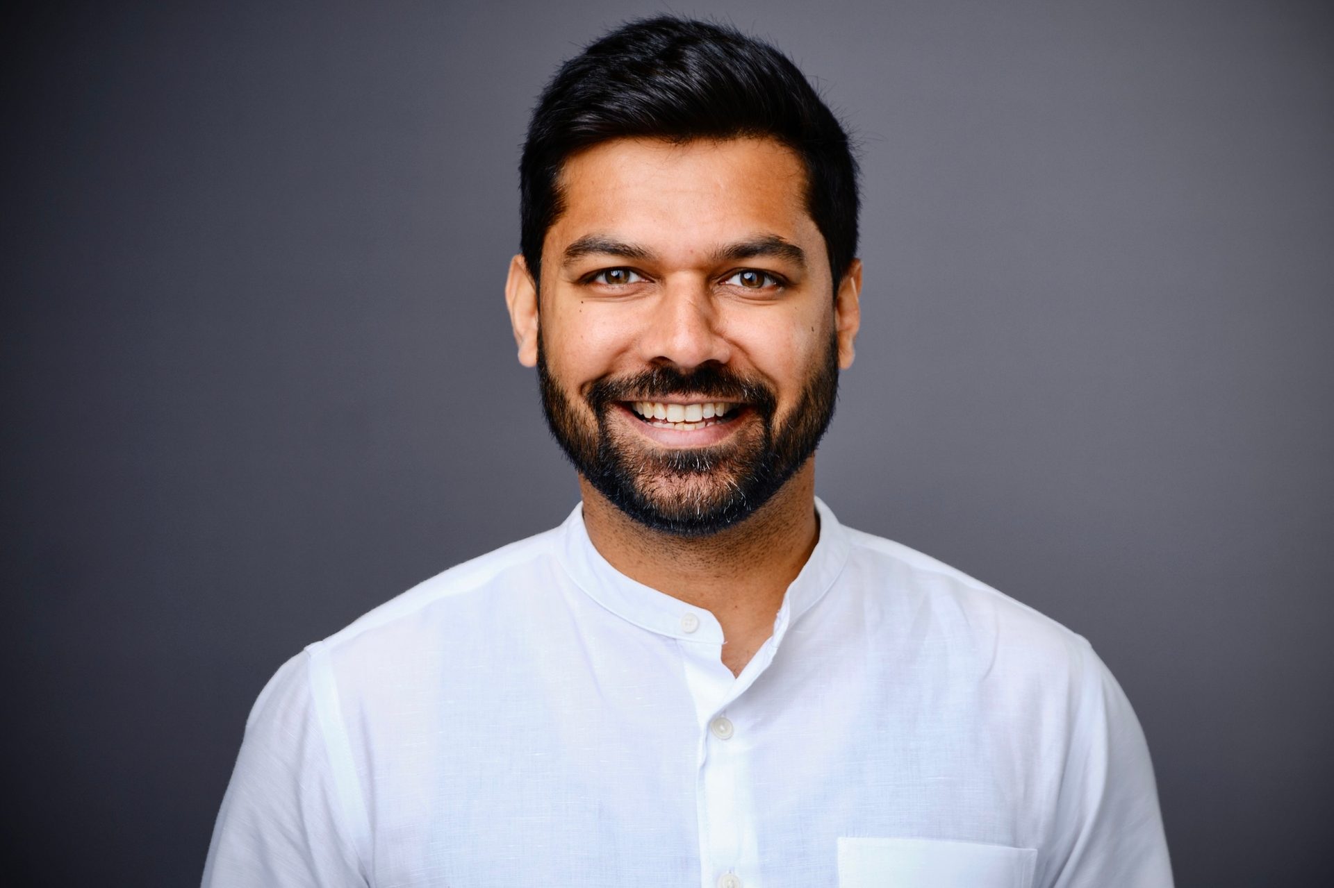 Headshot of a smiling man with a beard and dark hair, wearing a white collared shirt against a gray background.