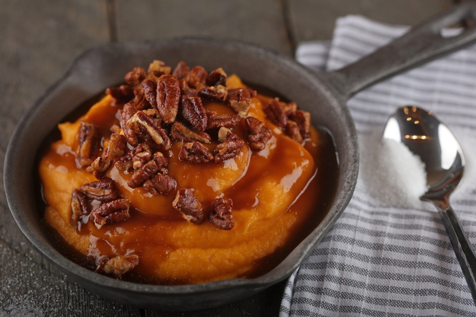 Sweet potato mash with pecans and caramel sauce in a skillet, next to a spoon with sugar.