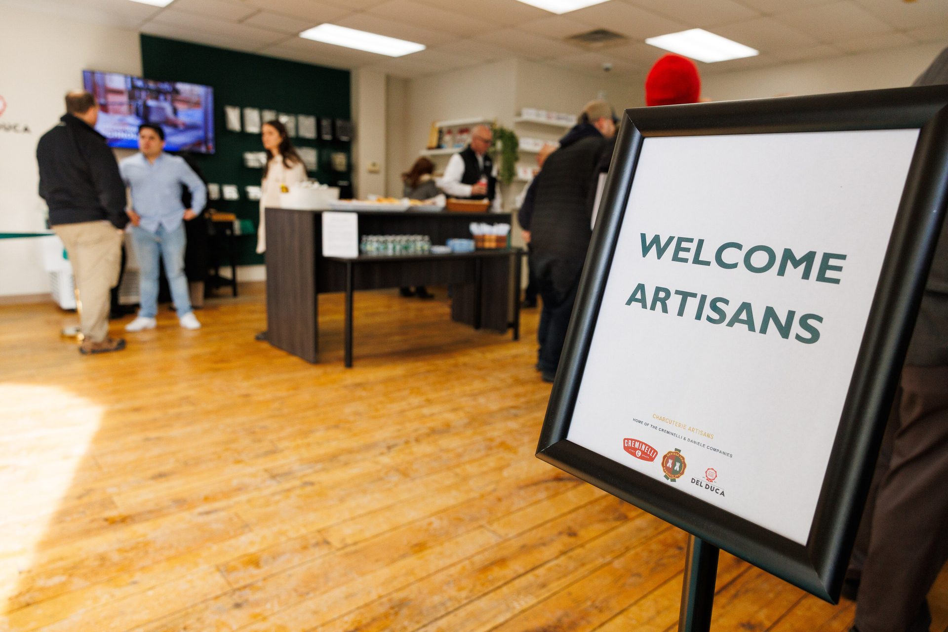 A 'Welcome Artisans' sign in the foreground, with people and a refreshment table in the background.