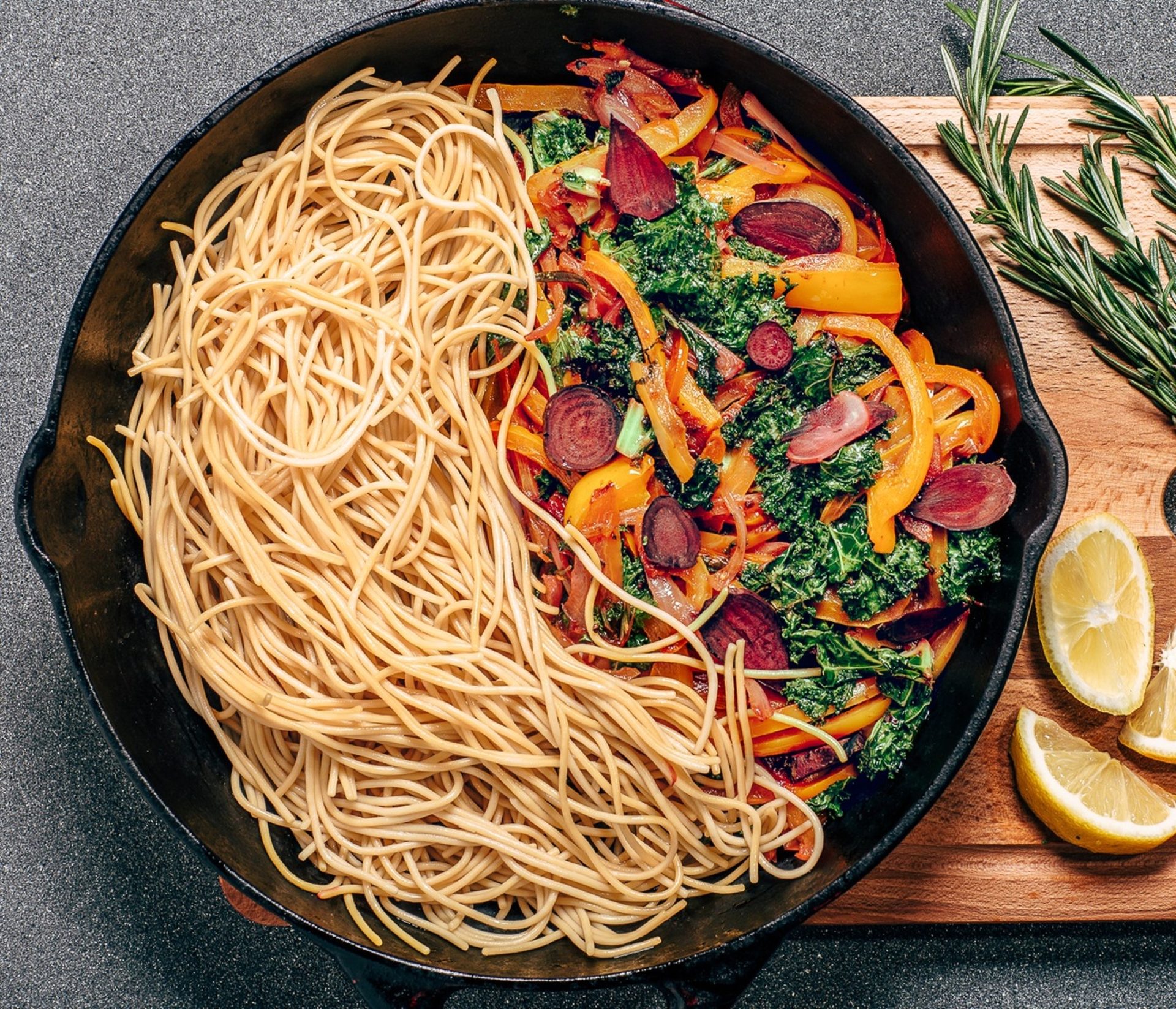 Skillet with spaghetti, sautéed vegetables (kale, peppers, beets), lemons, and rosemary.