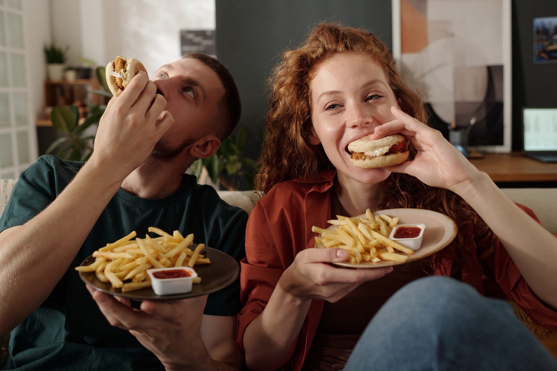 A man and a woman on a couch enjoying fast food: burgers/sandwiches and french fries with ketchup.