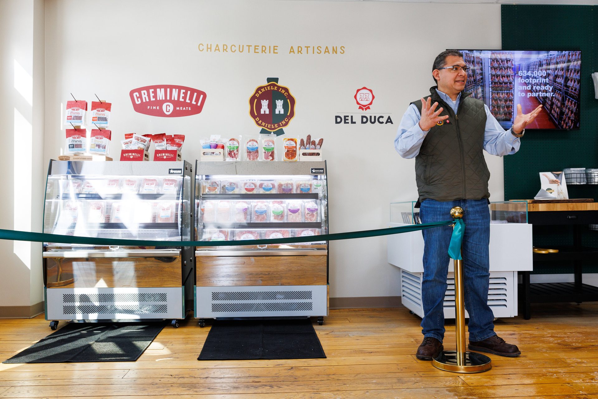 A man speaks in front of charcuterie display cases and wall branding for Cremninelli, Daniele Inc, and Del Duca.