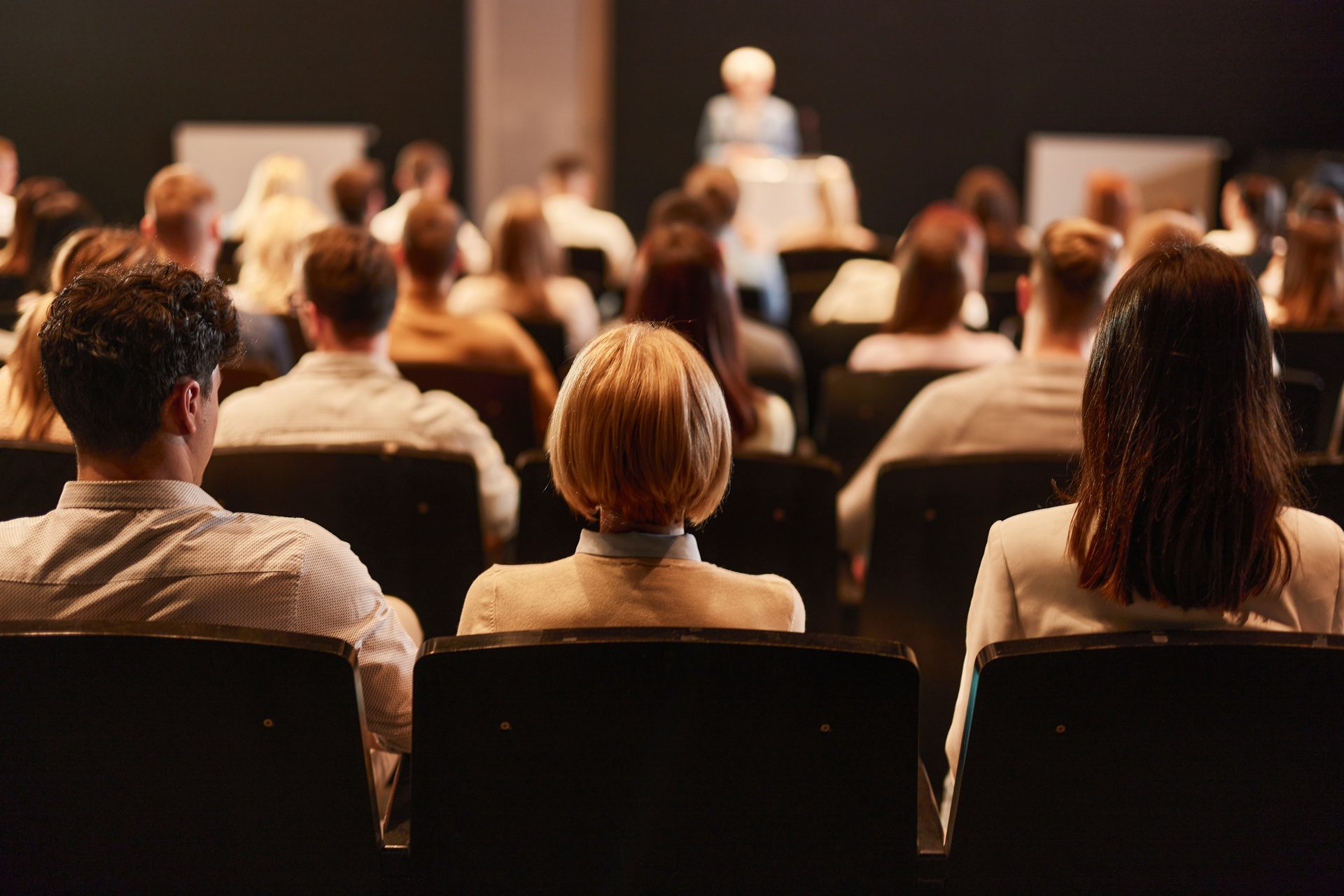 Audience backs facing a speaker on stage.