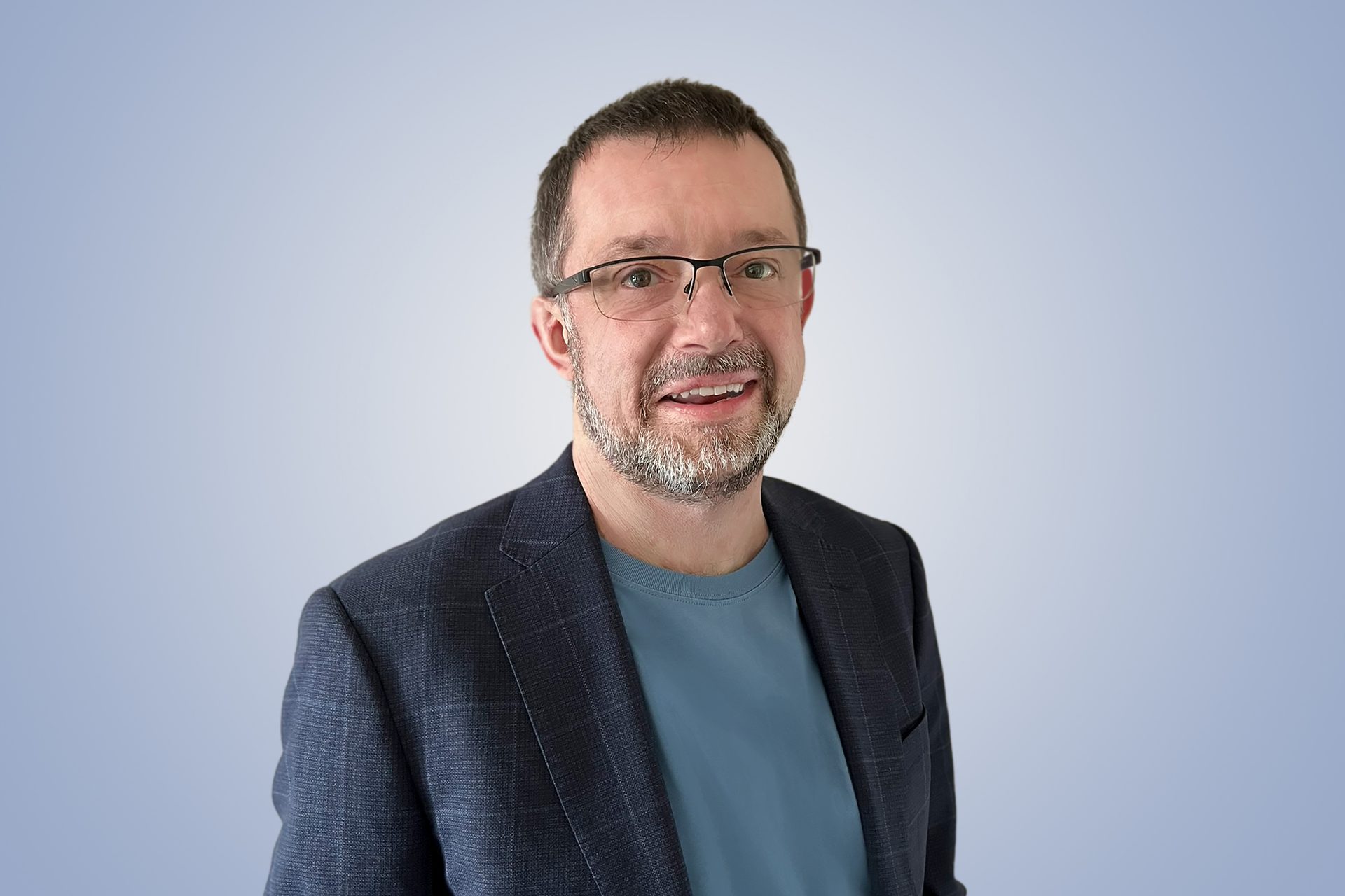 Smiling man with glasses, gray beard, dark jacket, and blue t-shirt against a light blue background.