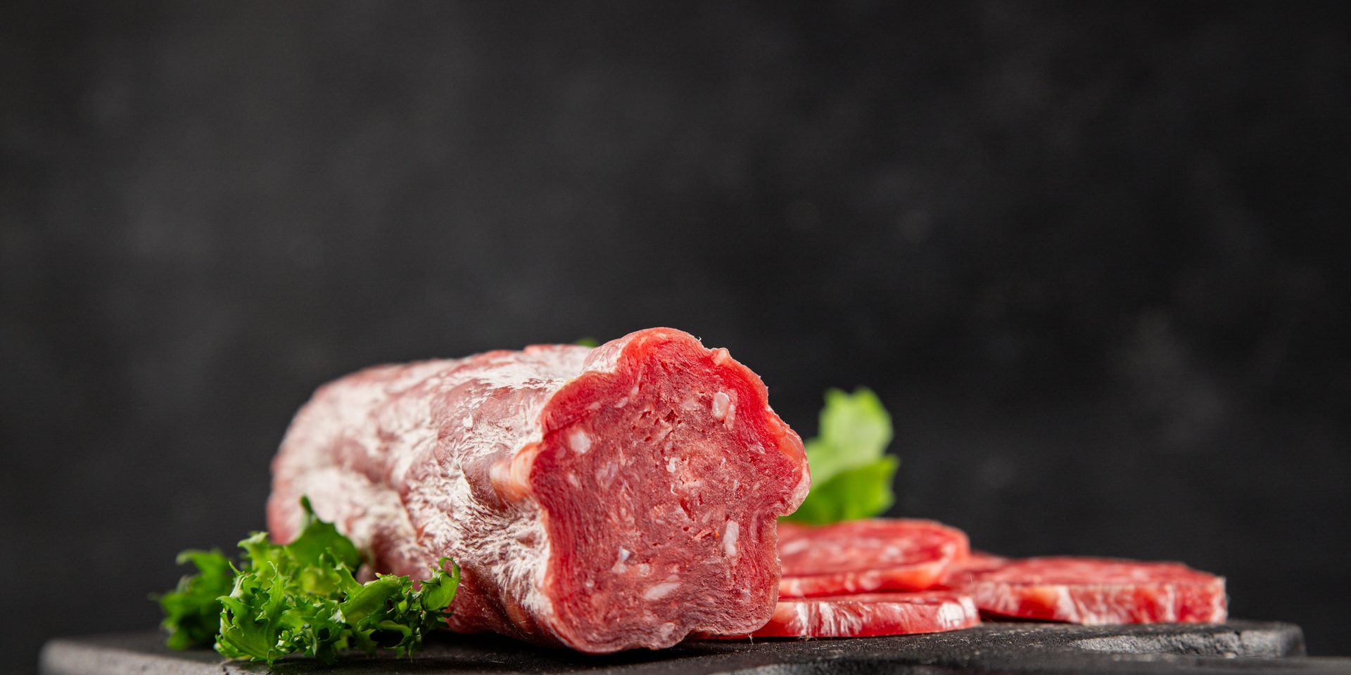 Partially sliced salami log with green garnish on a dark background.