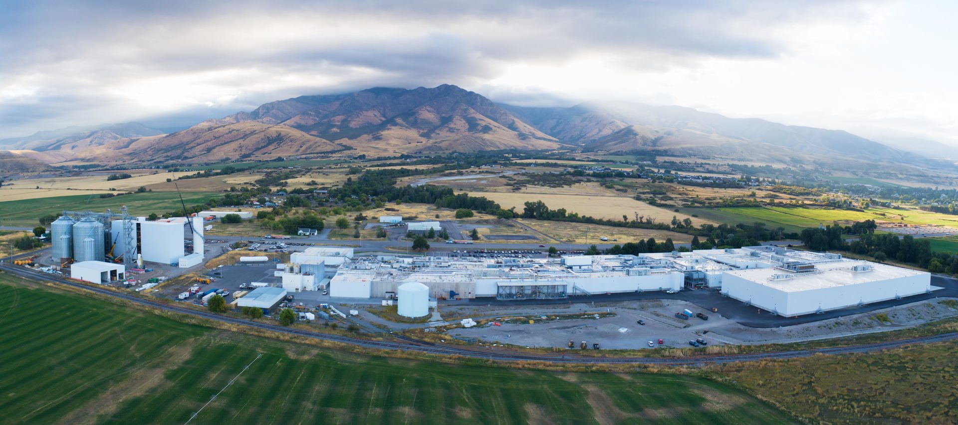 Aerial view of a large factory, silos, fields, and mountains under a dramatic sky.